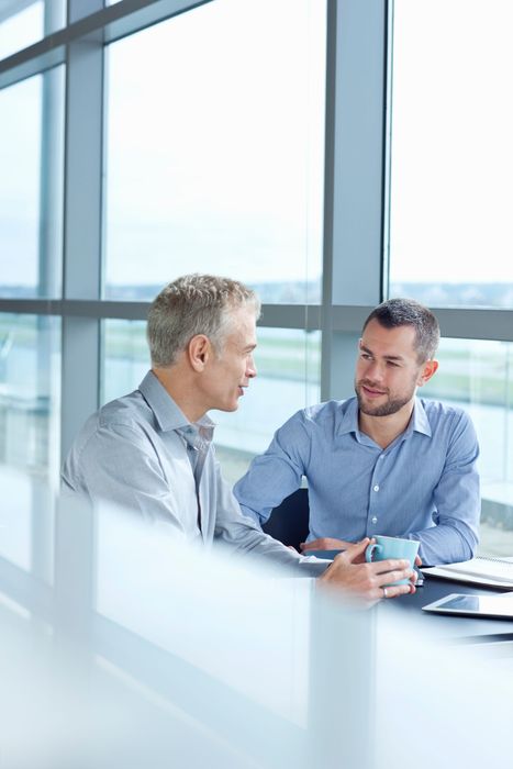Two men in a bright office meeting, discussing papers at a table with large windows behind them