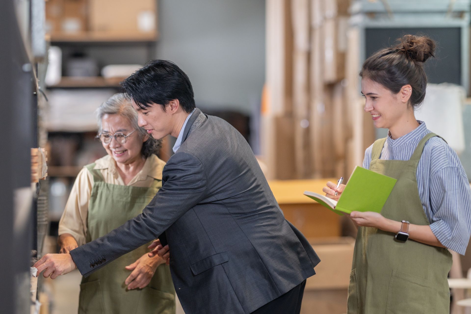 Three people in aprons in a workshop, one showing another how to use equipment while a third holds a clipboard.