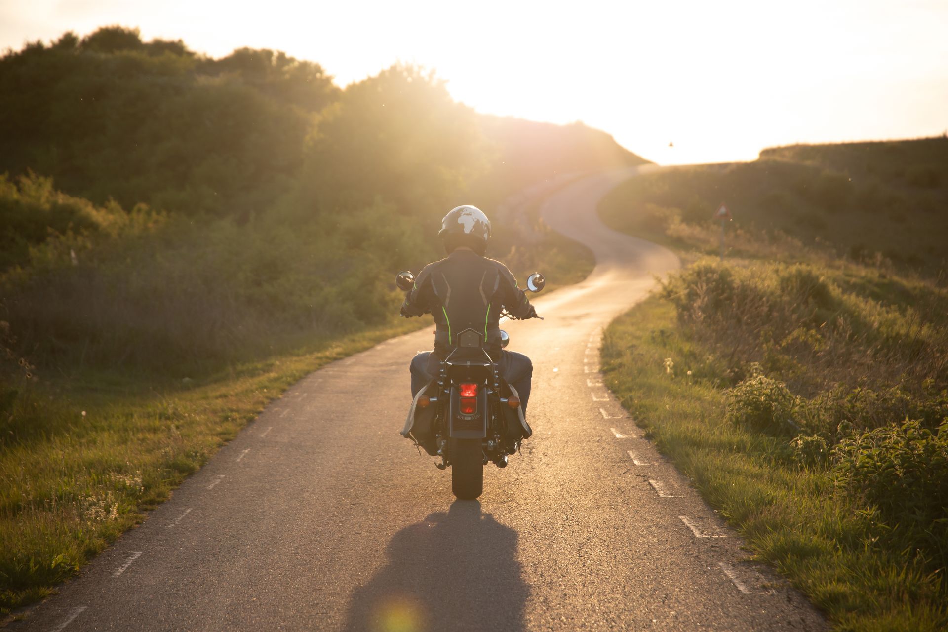 Motorcyclist rides a sunlit country road at sunset, with hills and greenery on both sides.