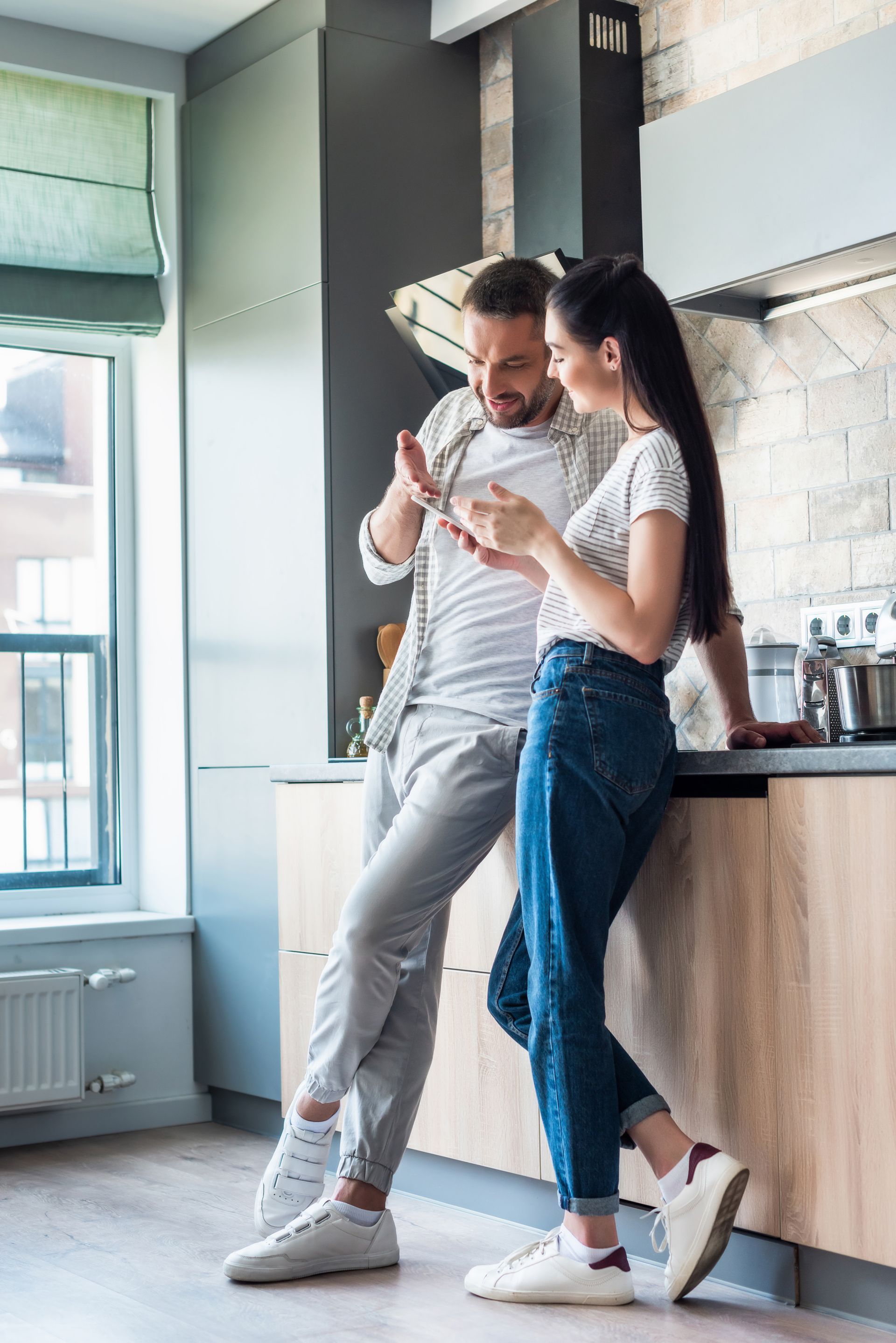 Couple leaning together in a bright kitchen, sharing a phone and coffee.