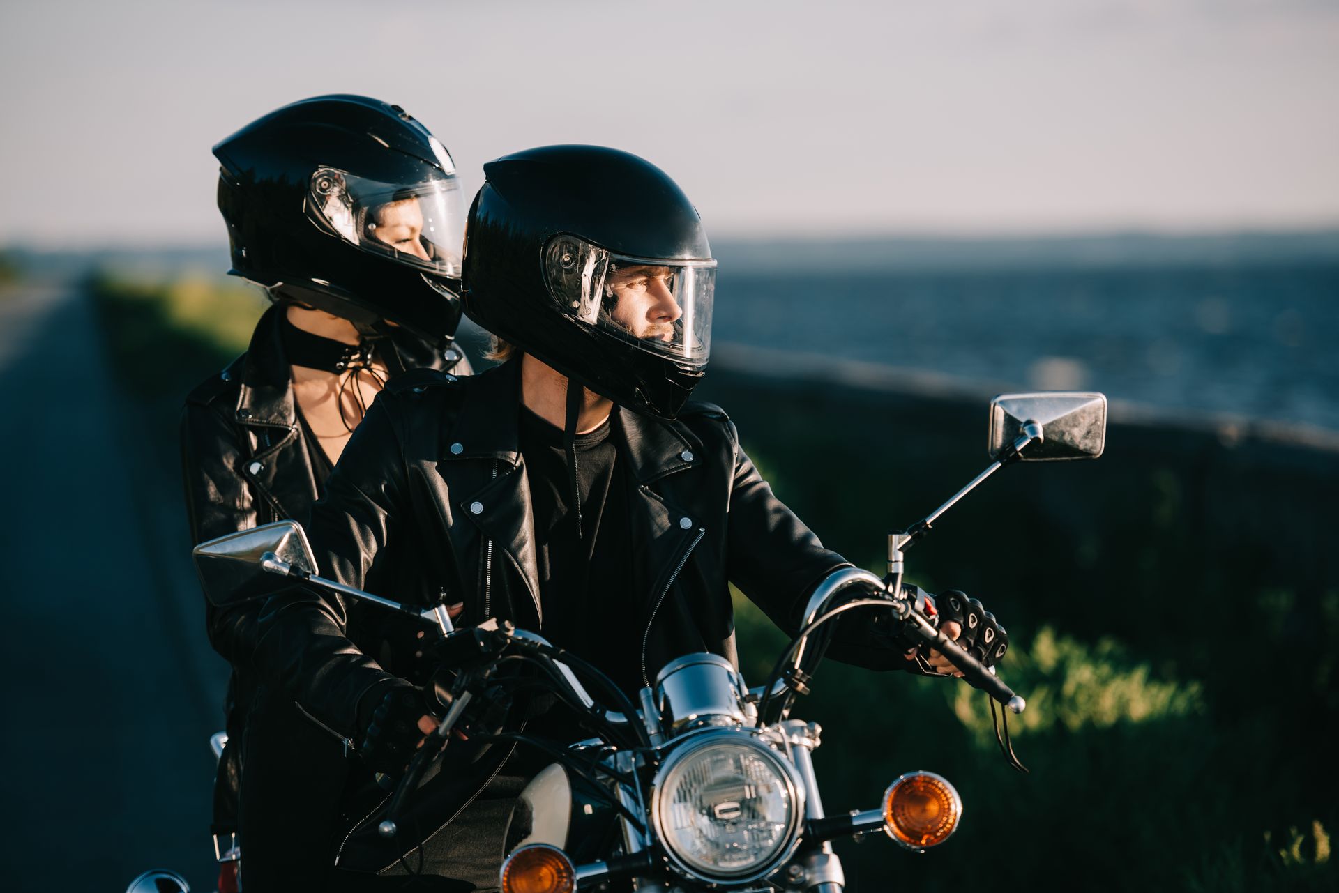 Two motorcyclists in black helmets and jackets riding at dusk with the ocean in the background