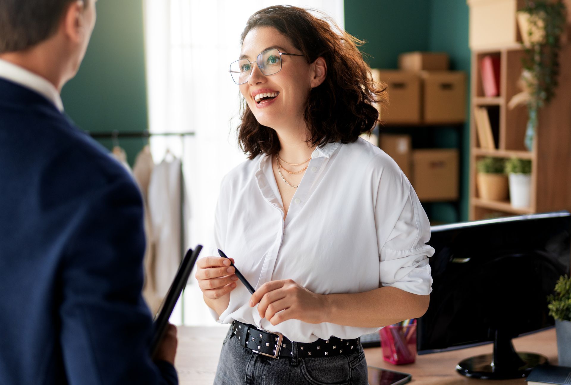 Two people talking in an office, with a woman in a white blouse holding glasses near a desk.