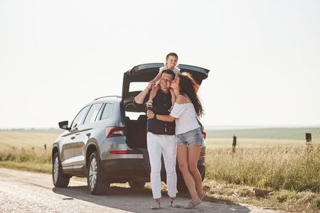 Family posing by an SUV with open trunk on a rural roadside, smiling in a sunny field