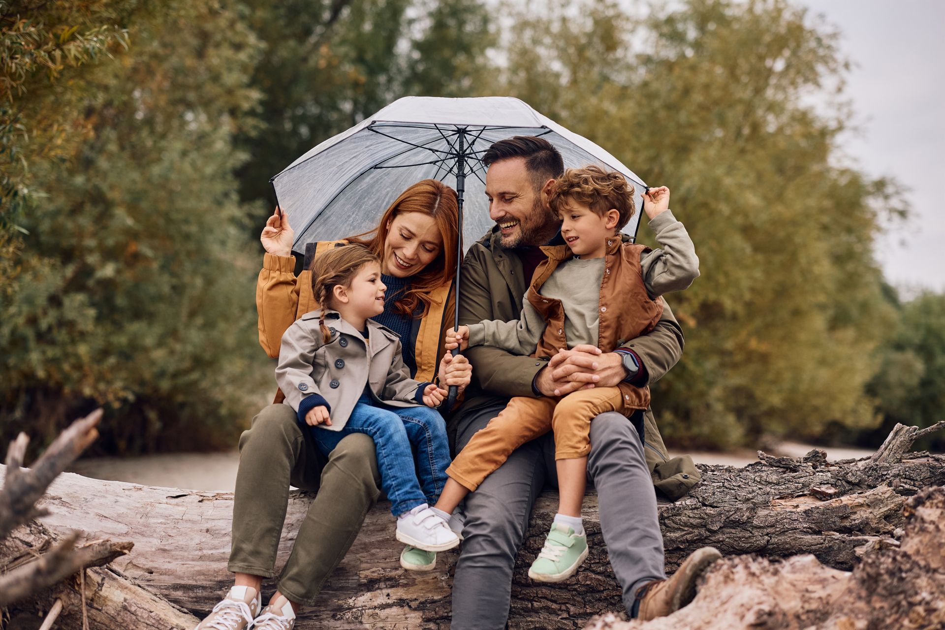 Family sitting together under an umbrella on a fallen log in a wooded outdoor setting
