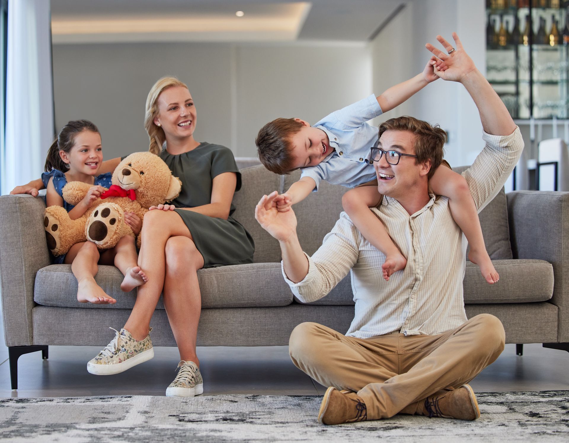 Family on a gray sofa and floor laughing together, with teddy bears in a bright living room