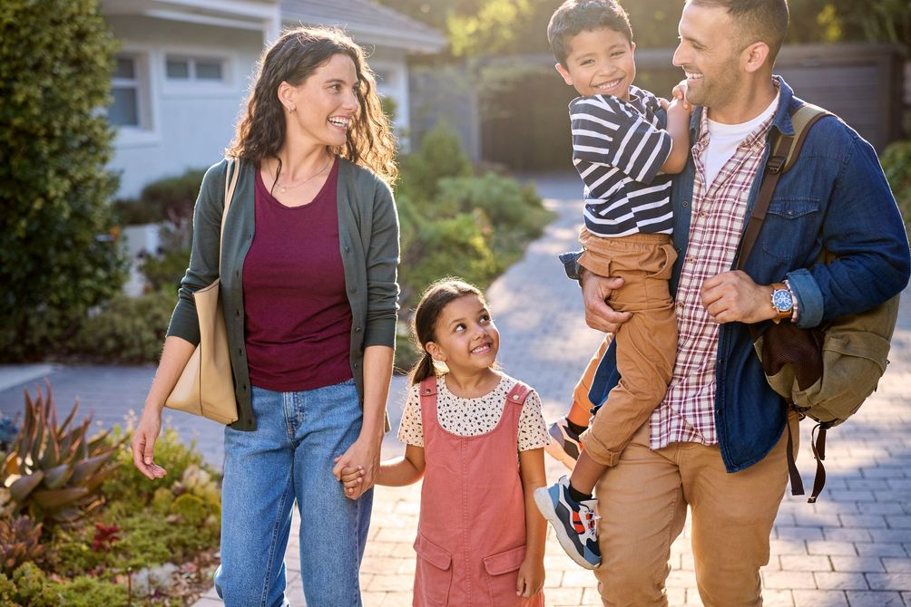 Family of four walking outdoors by a house, smiling and holding hands in warm sunlight
