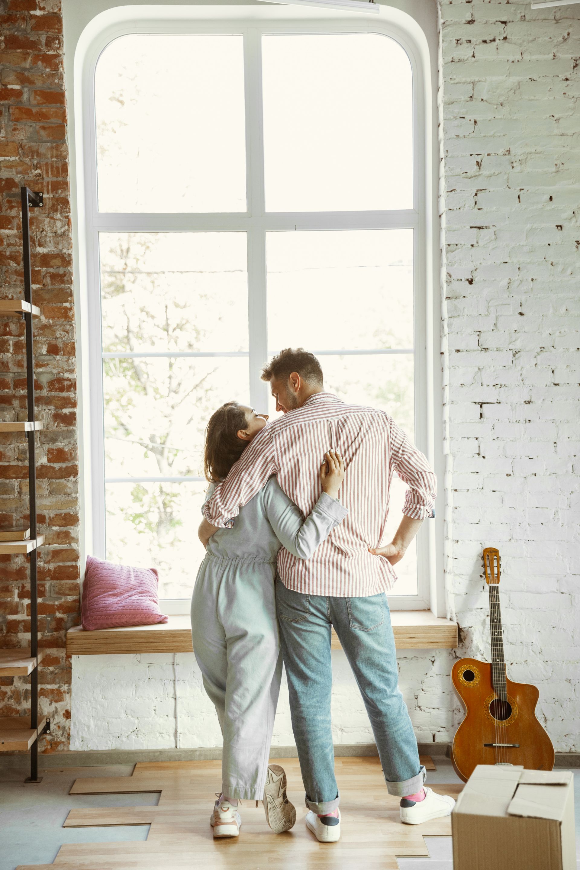 Couple standing by a large bright window in a cozy studio with a guitar and bench nearby