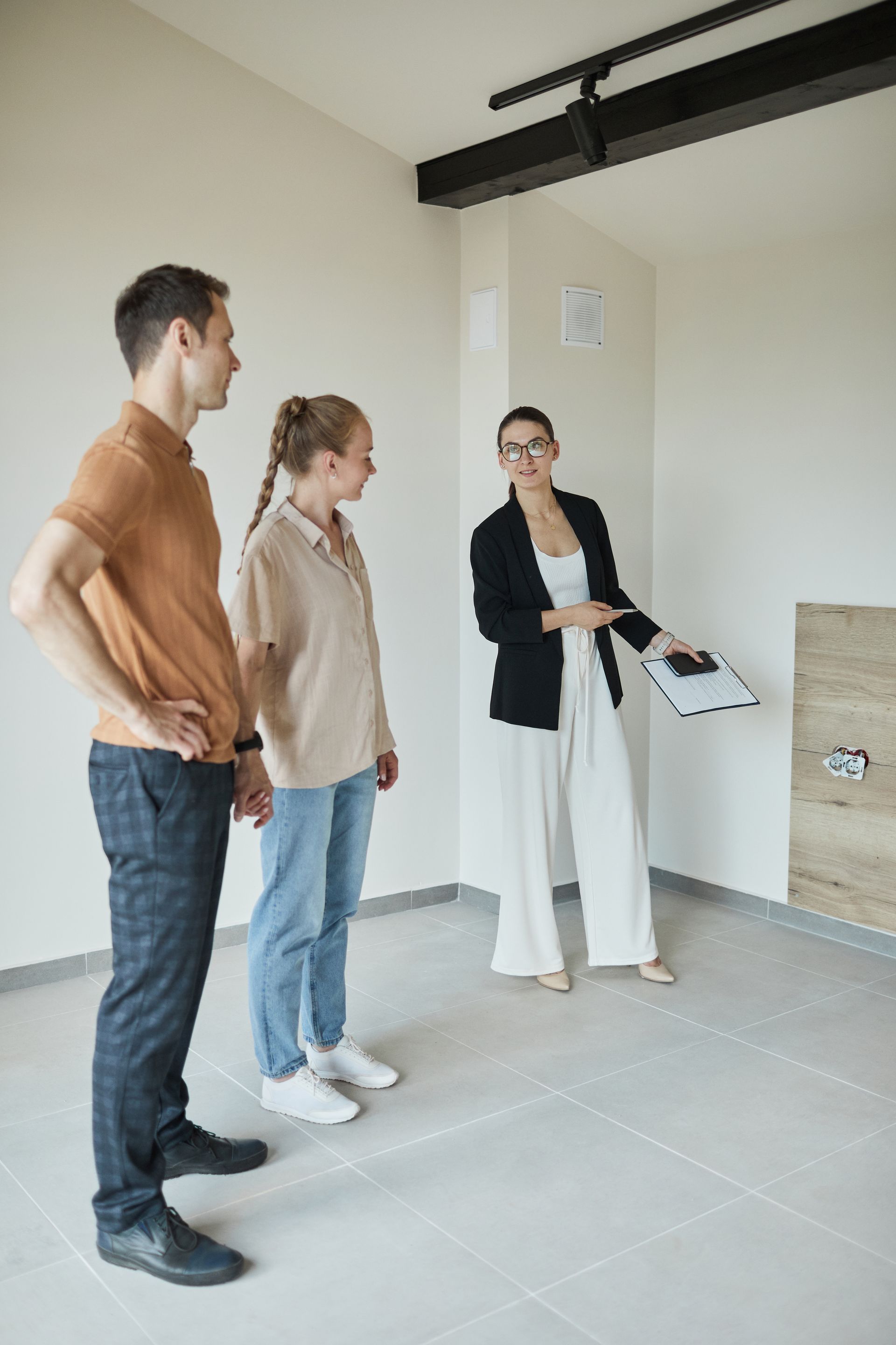 Three people stand in an unfinished room, looking at a person holding papers and gesturing.