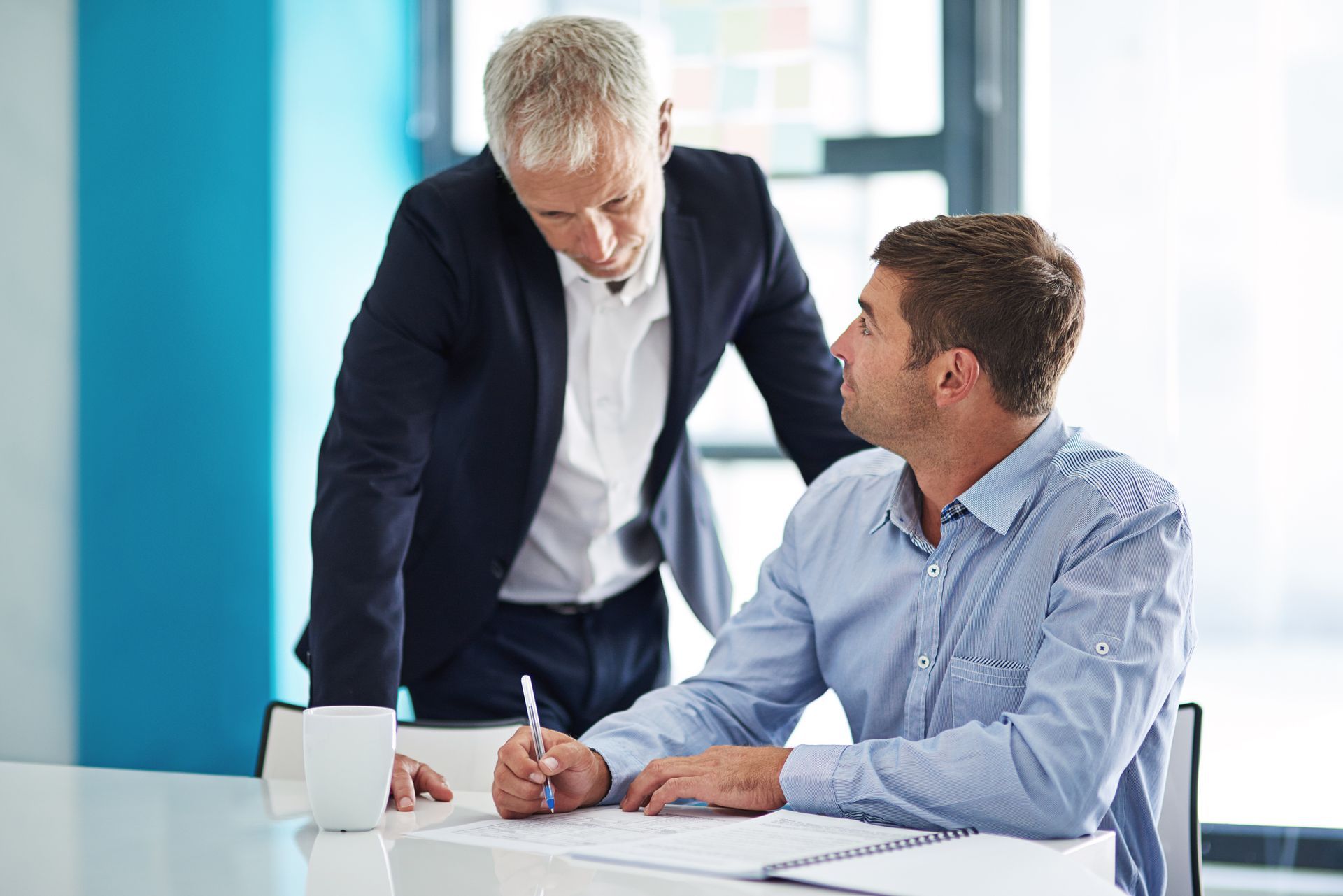 Two coworkers discuss a document at a desk in a bright office.