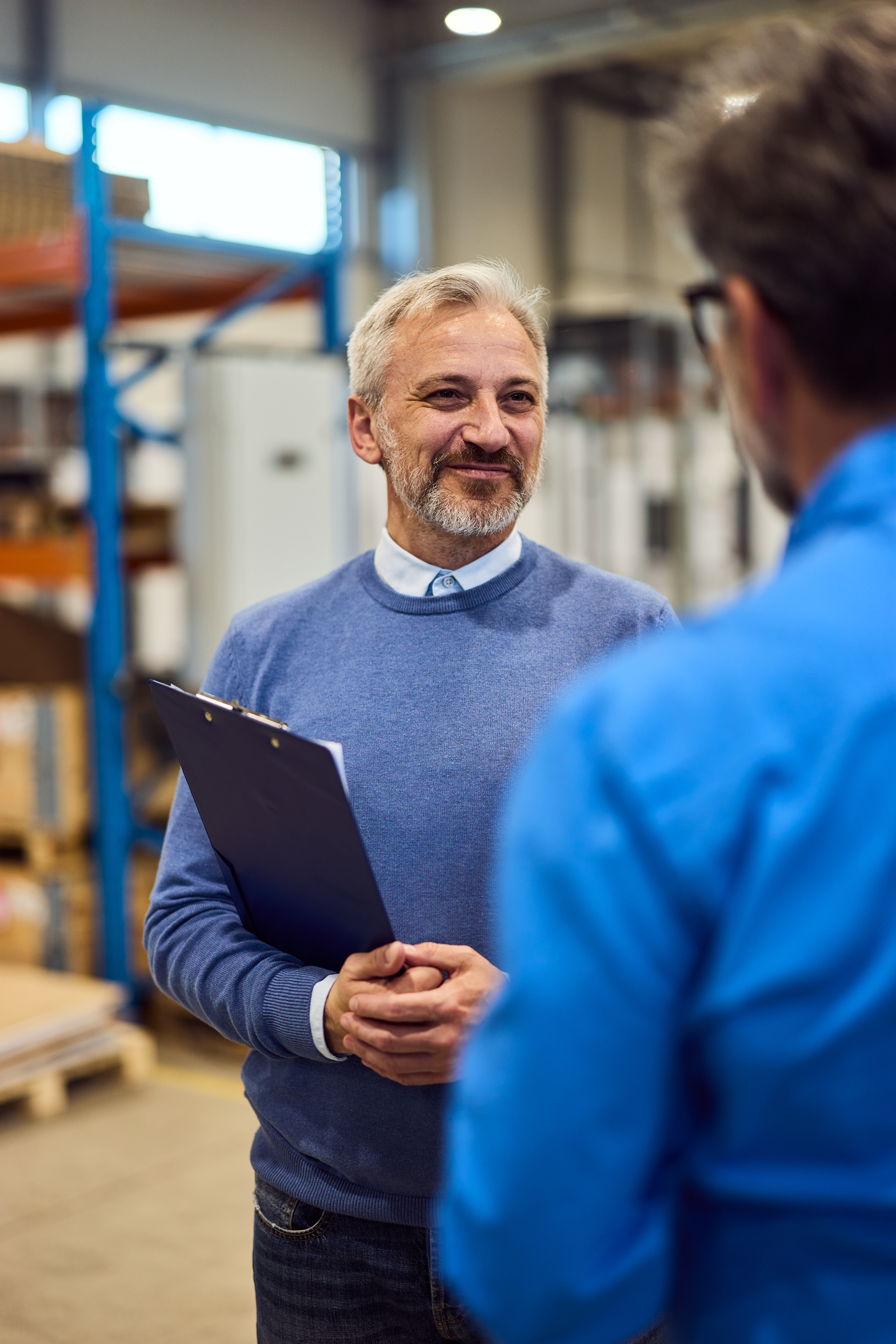 Two people talking in a warehouse, one holding a clipboard and smiling.