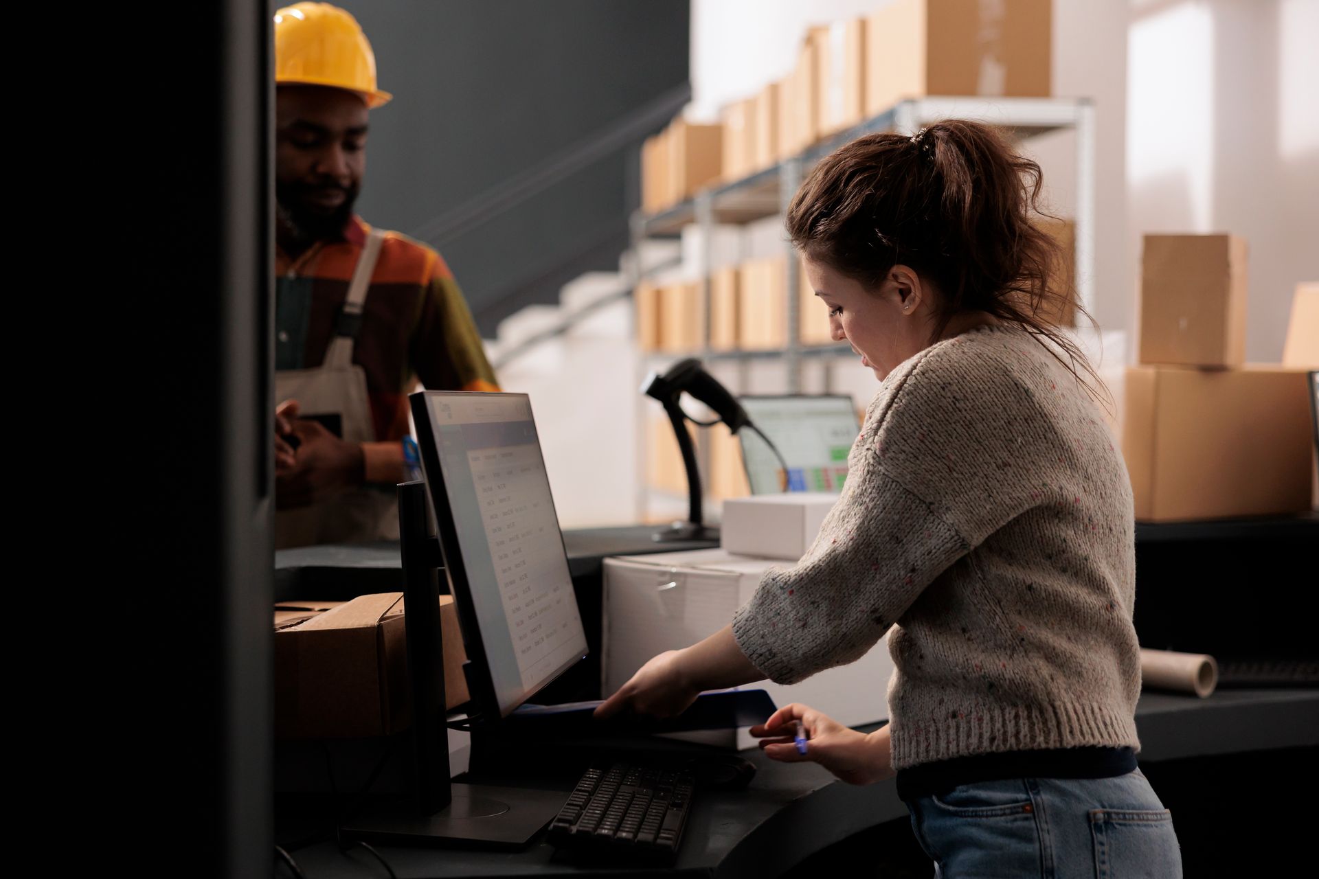 Person at a computer in a warehouse, with stacked cardboard boxes and a worker in a hard hat nearby.