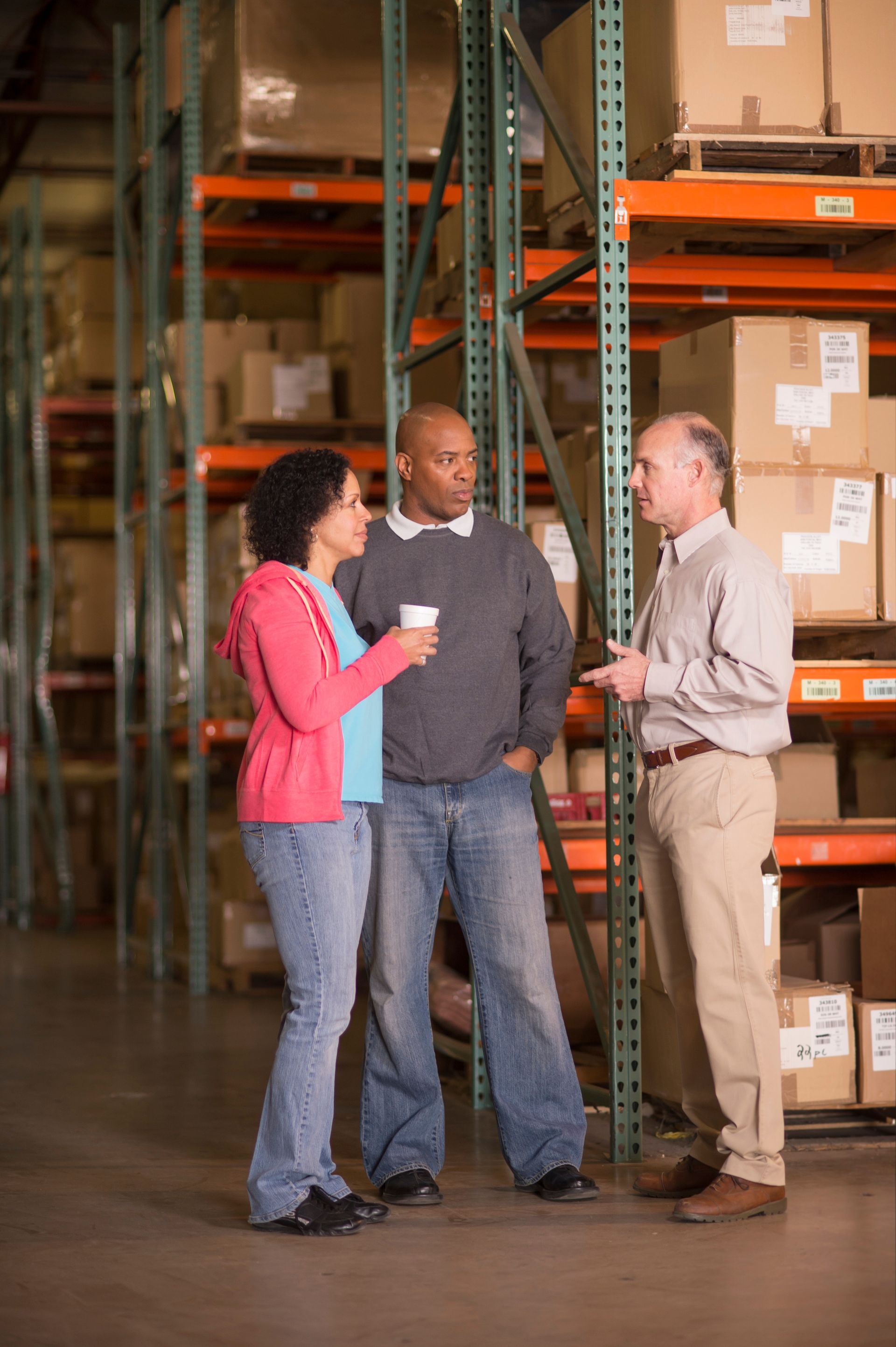 Three people talk in a warehouse aisle among stacked boxes and orange shelves.