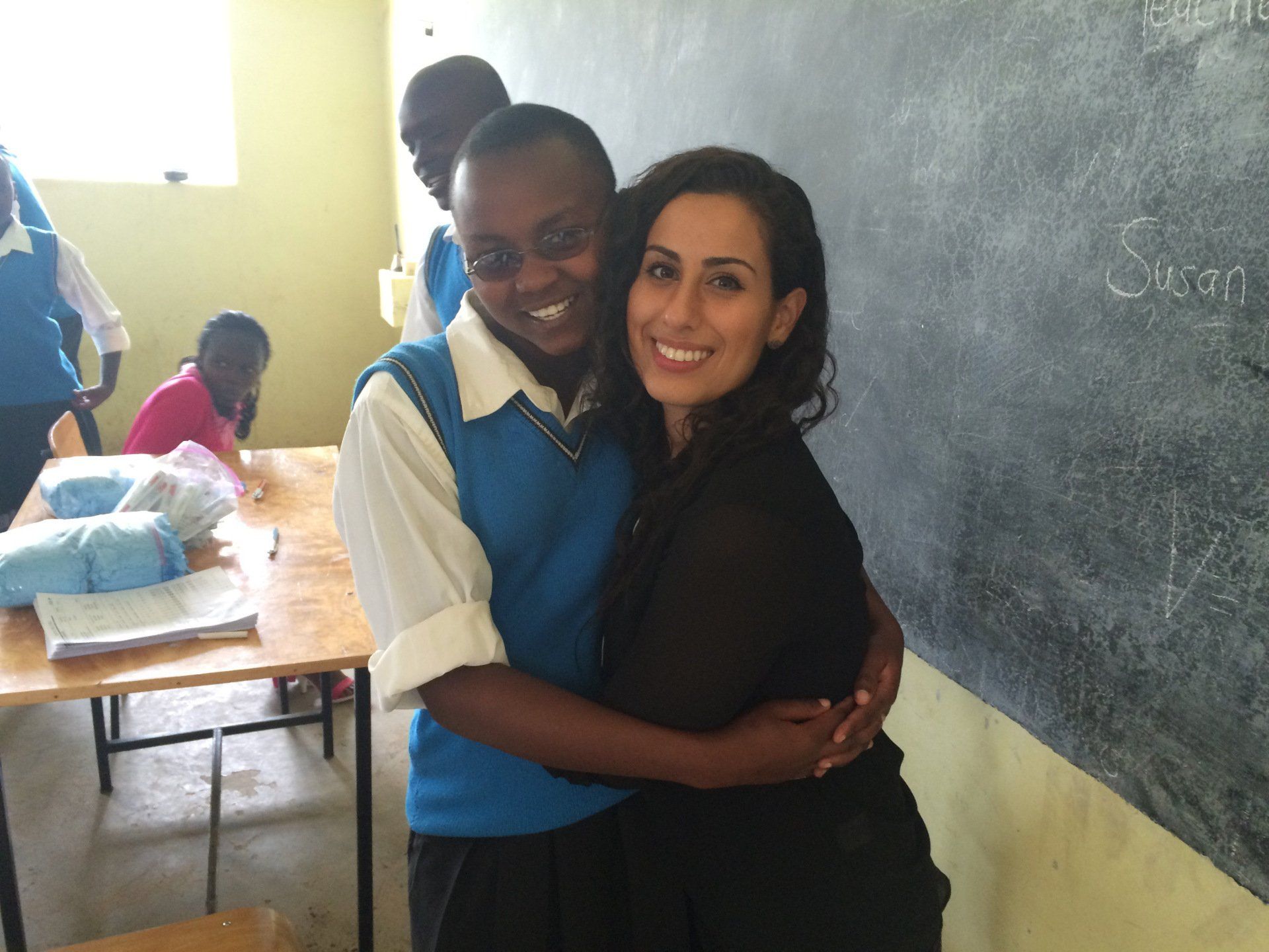A girl in a blue vest is hugging a woman in a classroom