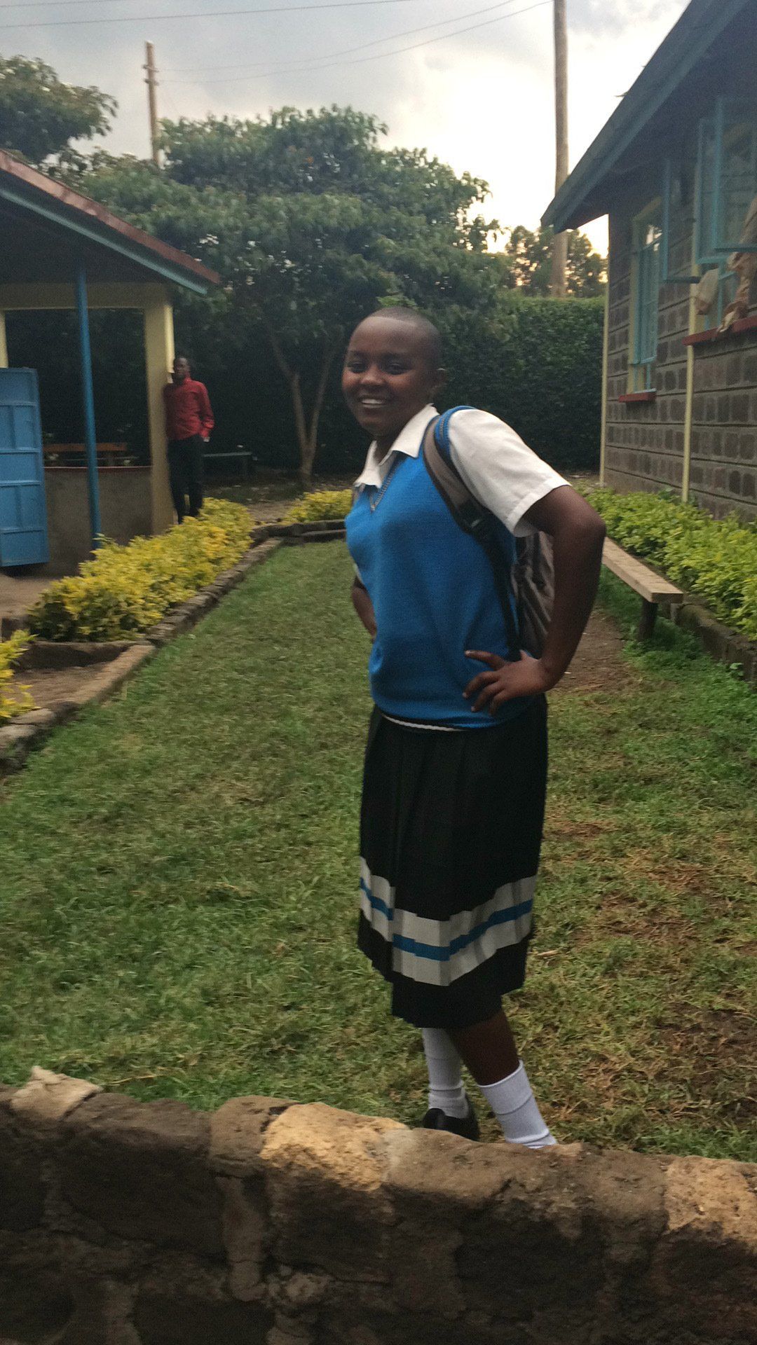 A young girl in a school uniform is standing in a grassy yard.