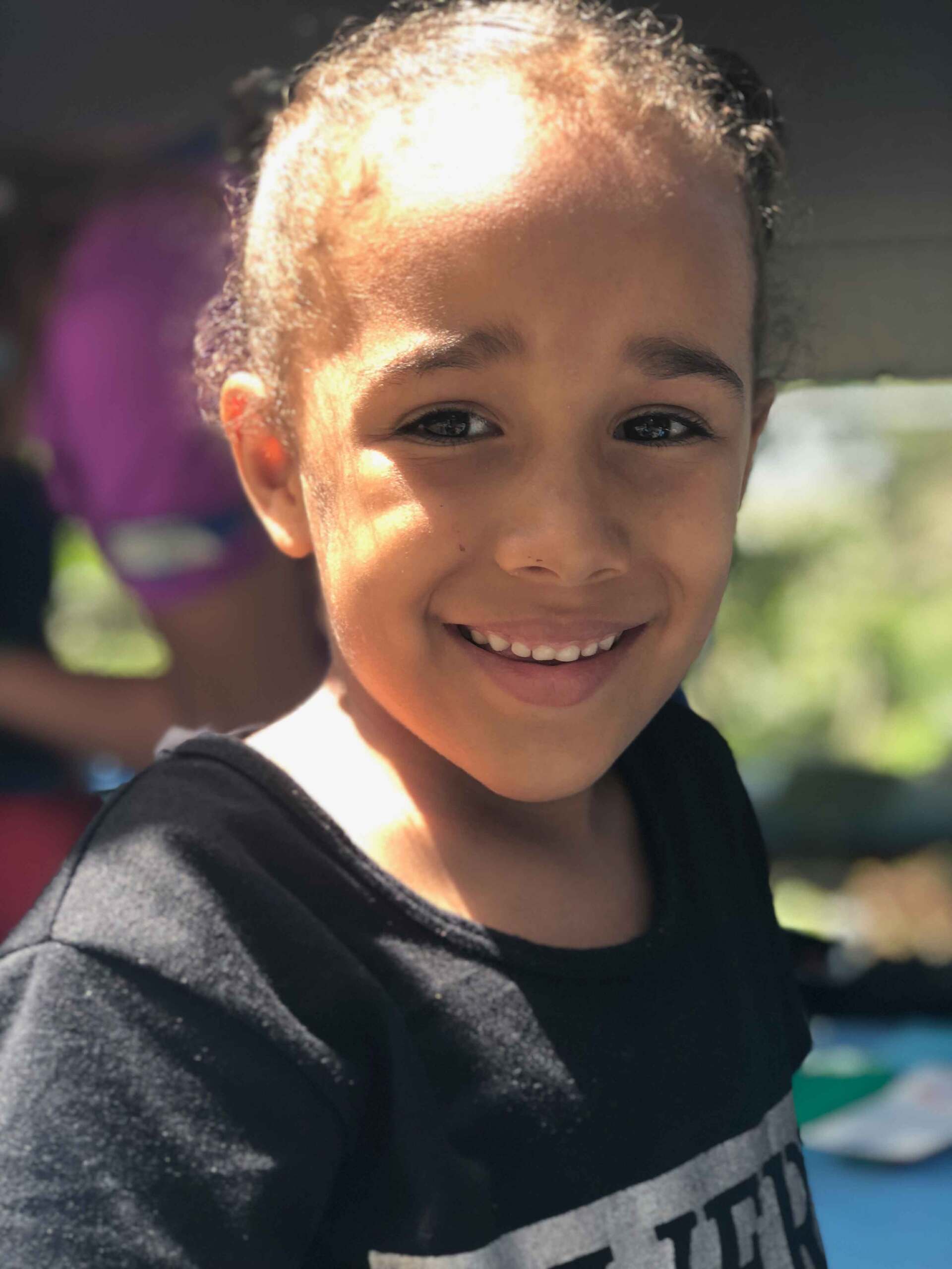 A young girl wearing a black shirt with the word tee on it smiles for the camera
