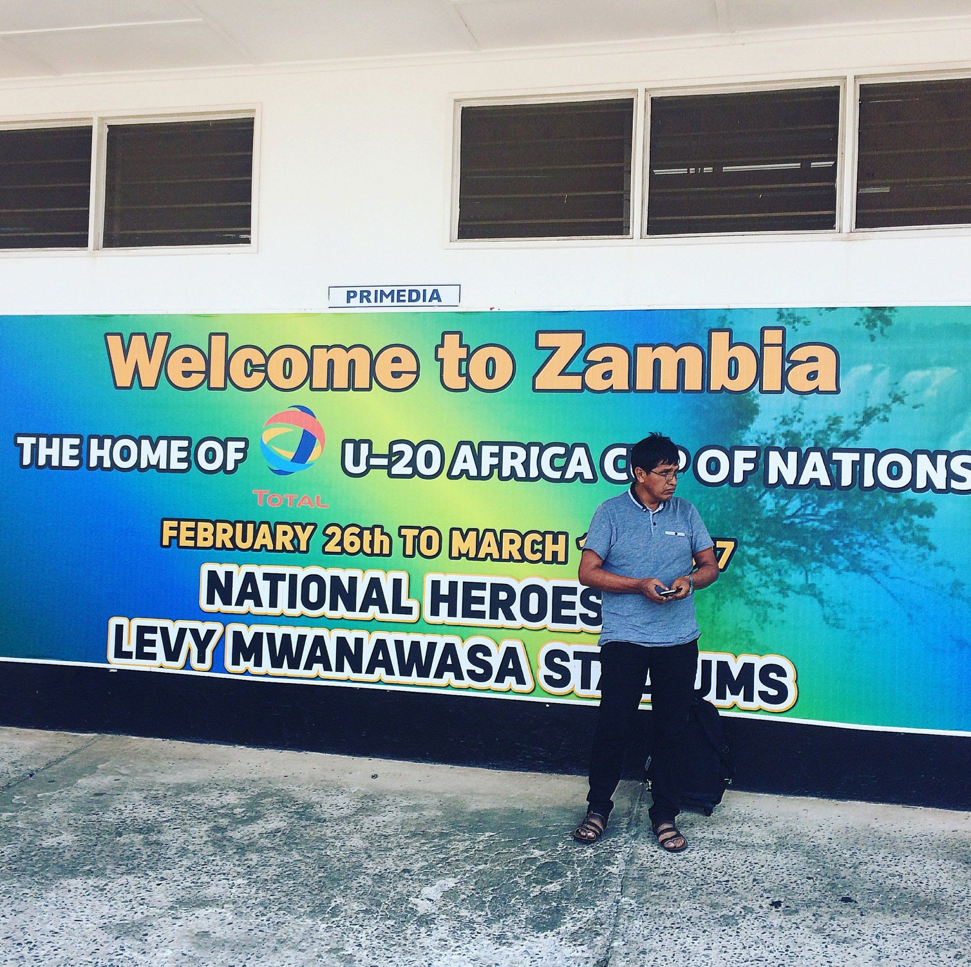 A man stands in front of a welcome to zambia sign