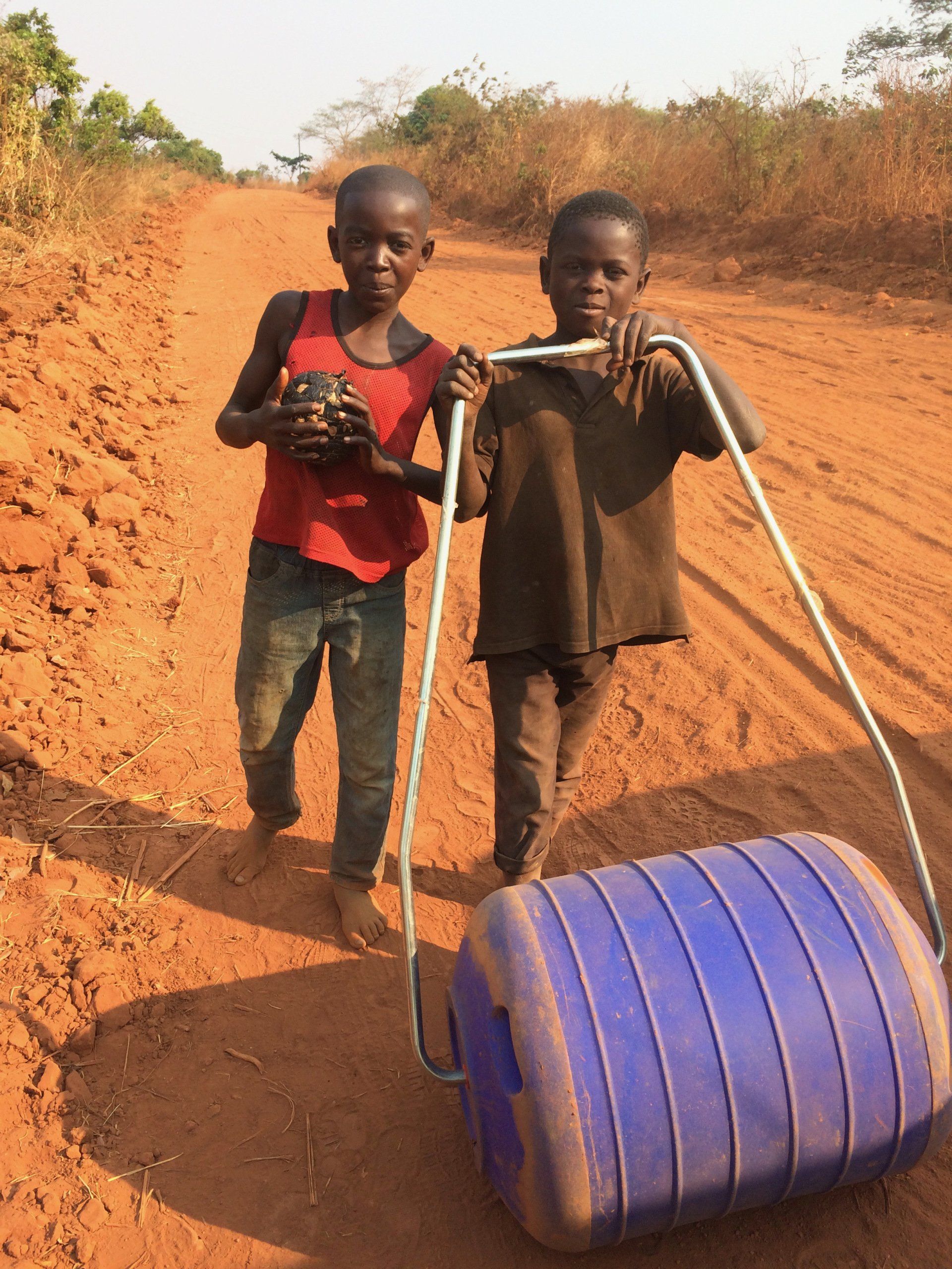 Two young boys standing next to a blue barrel on a dirt road