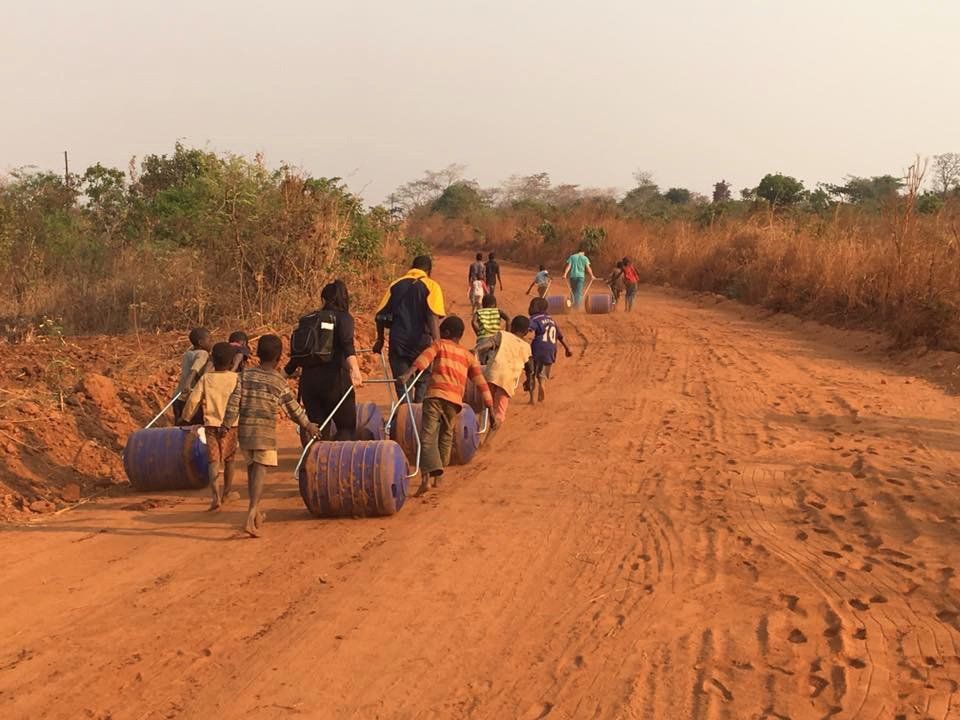 A group of people are walking down a dirt road carrying barrels.