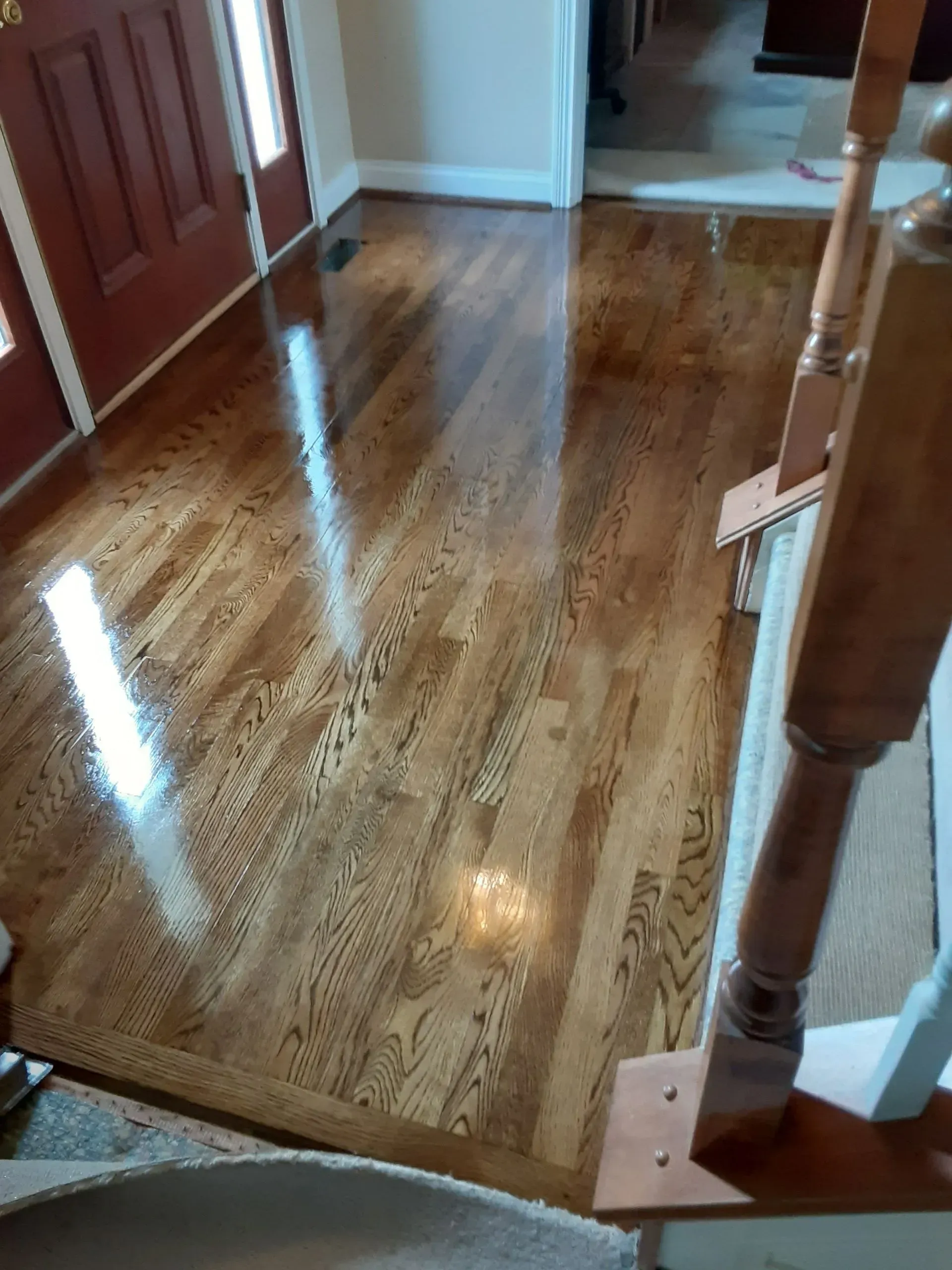 Polished wood floor in a home entryway, reflecting light. Staircase railing visible on right side. Red door on left.