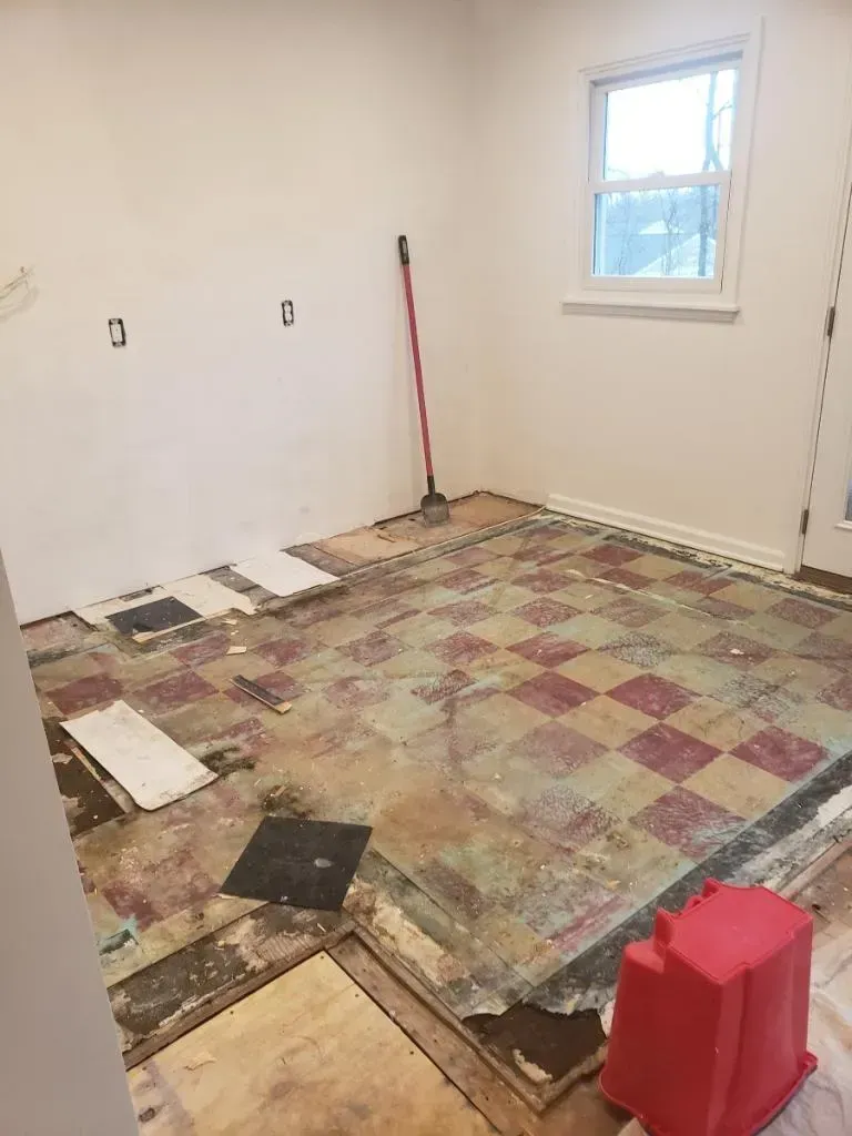 Room under renovation with damaged checkered floor, bare walls, and a window. A red bucket sits on the right.