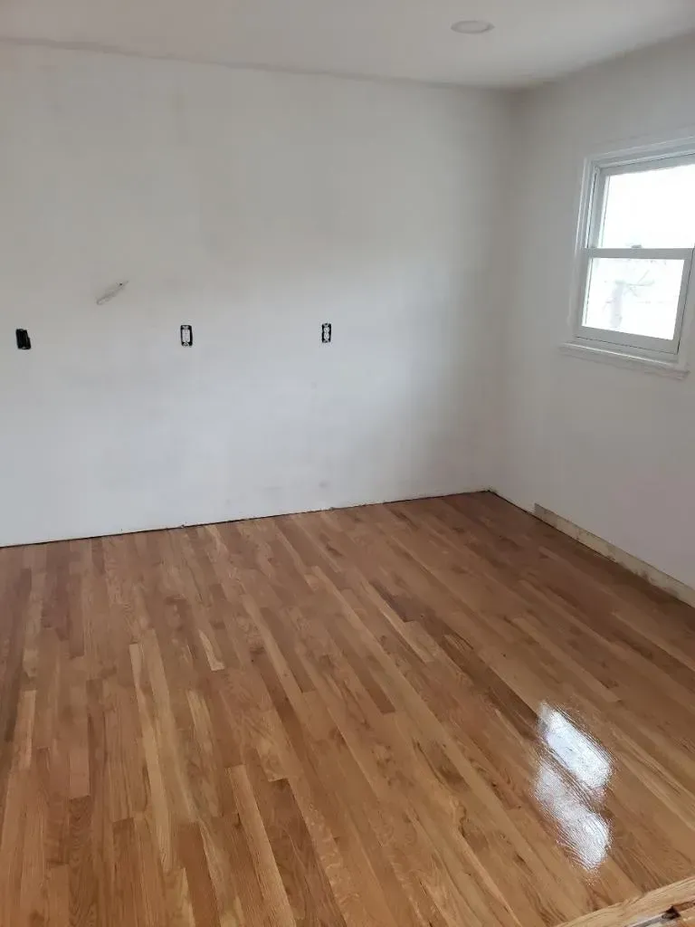 Empty room with newly installed hardwood floors, white walls, and a window.