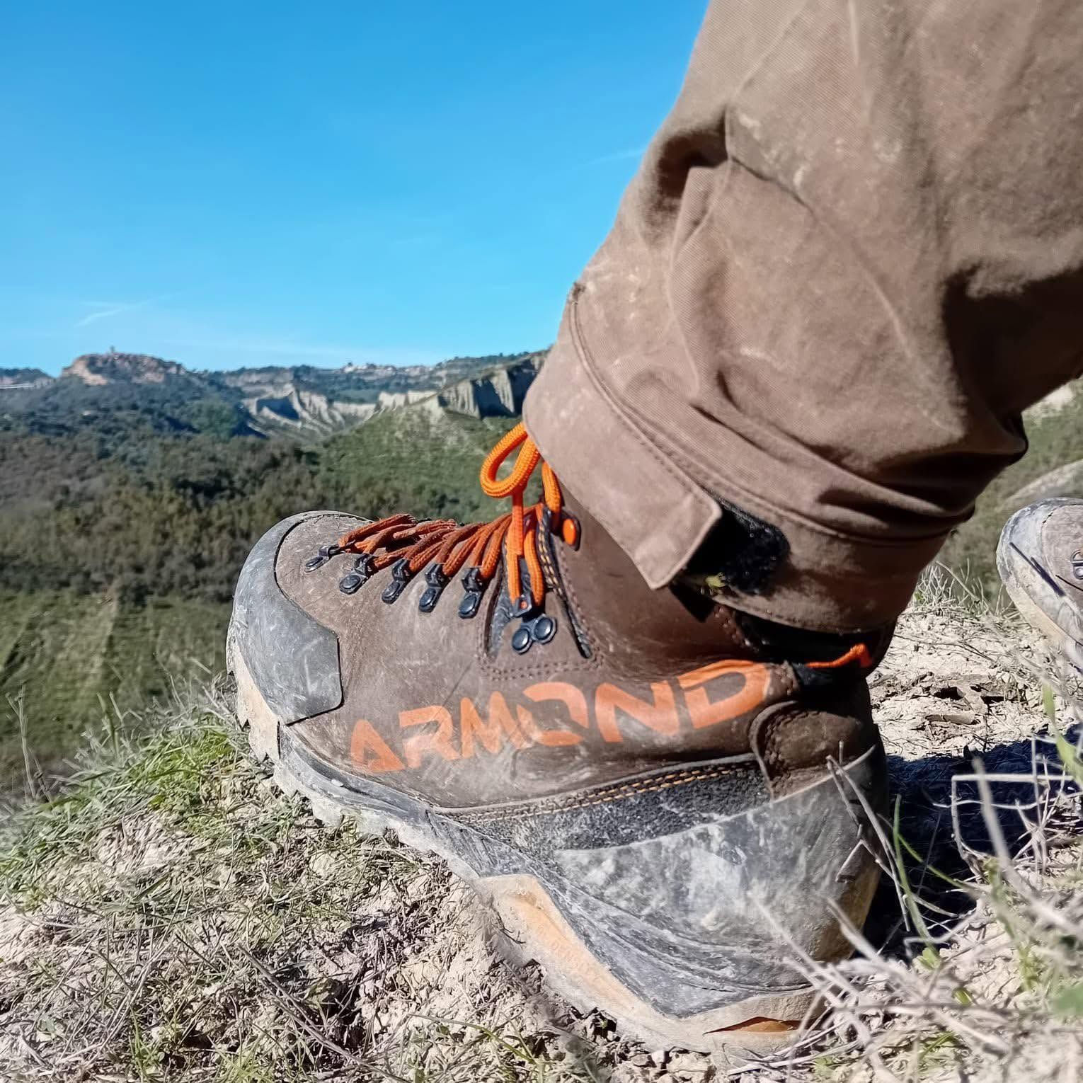 Primo piano di uno scarpone da trekking che cammina su un sentiero roccioso con montagne e cielo azzurro sullo sfondo.