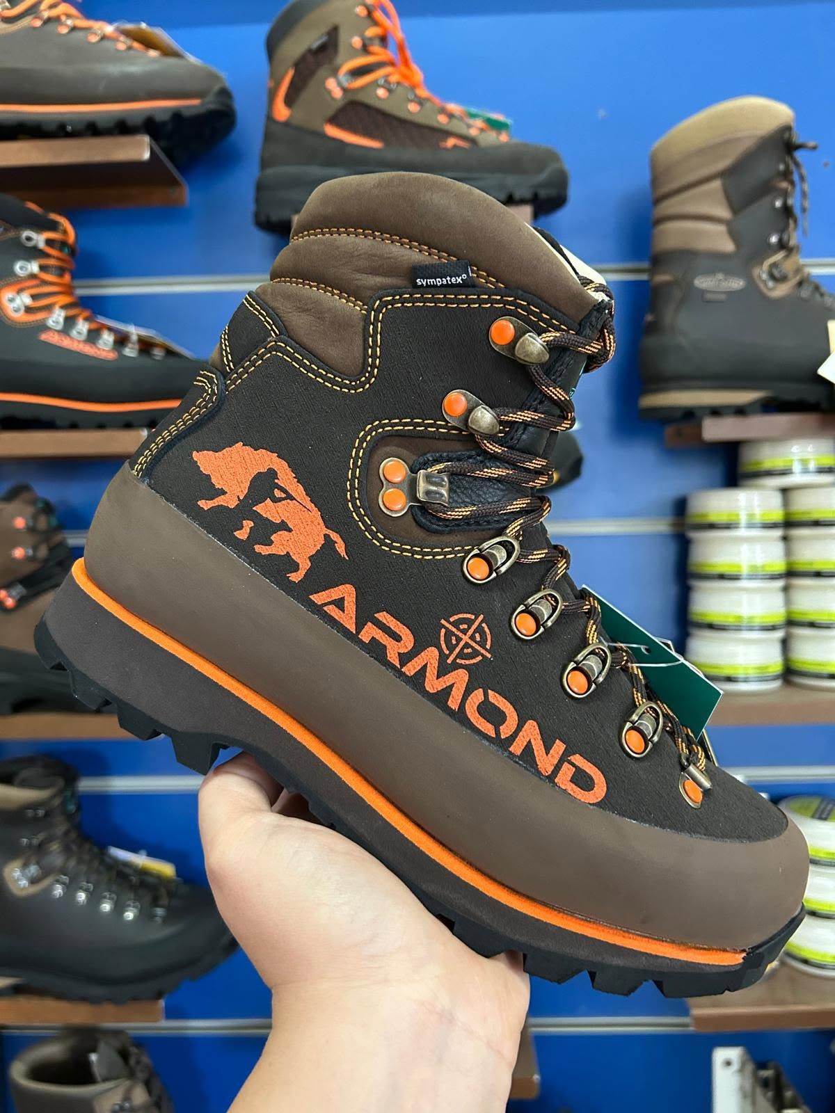 Uno scarpone da trekking ARMOND nero e arancione, tenuto in mano ed esposto in un negozio insieme ad altri scarponi.