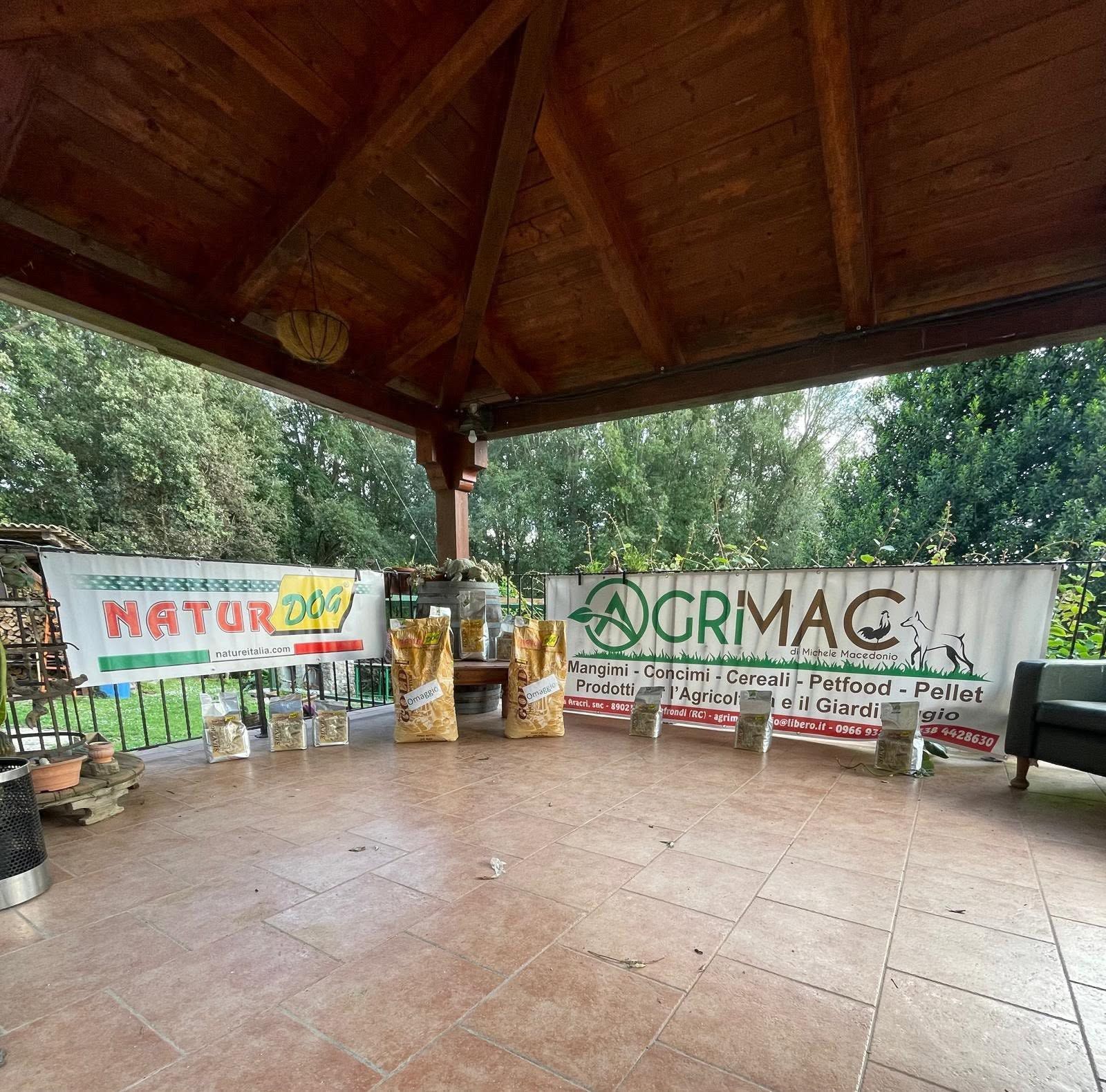 Stand esterno coperto con striscioni NATURA e QRMA, cesti di prodotti freschi e pavimento piastrellato.