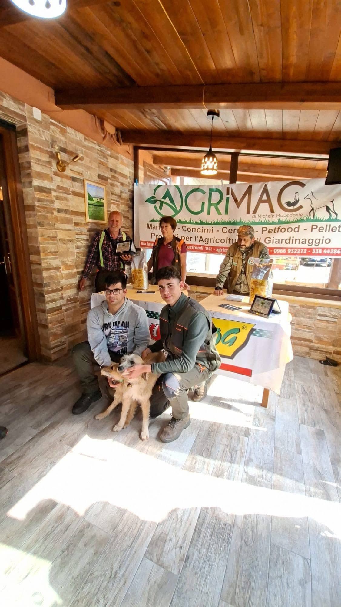 Tre persone con un cane presso una bancarella rustica di un mercato coperto con un cartello verde con la scritta