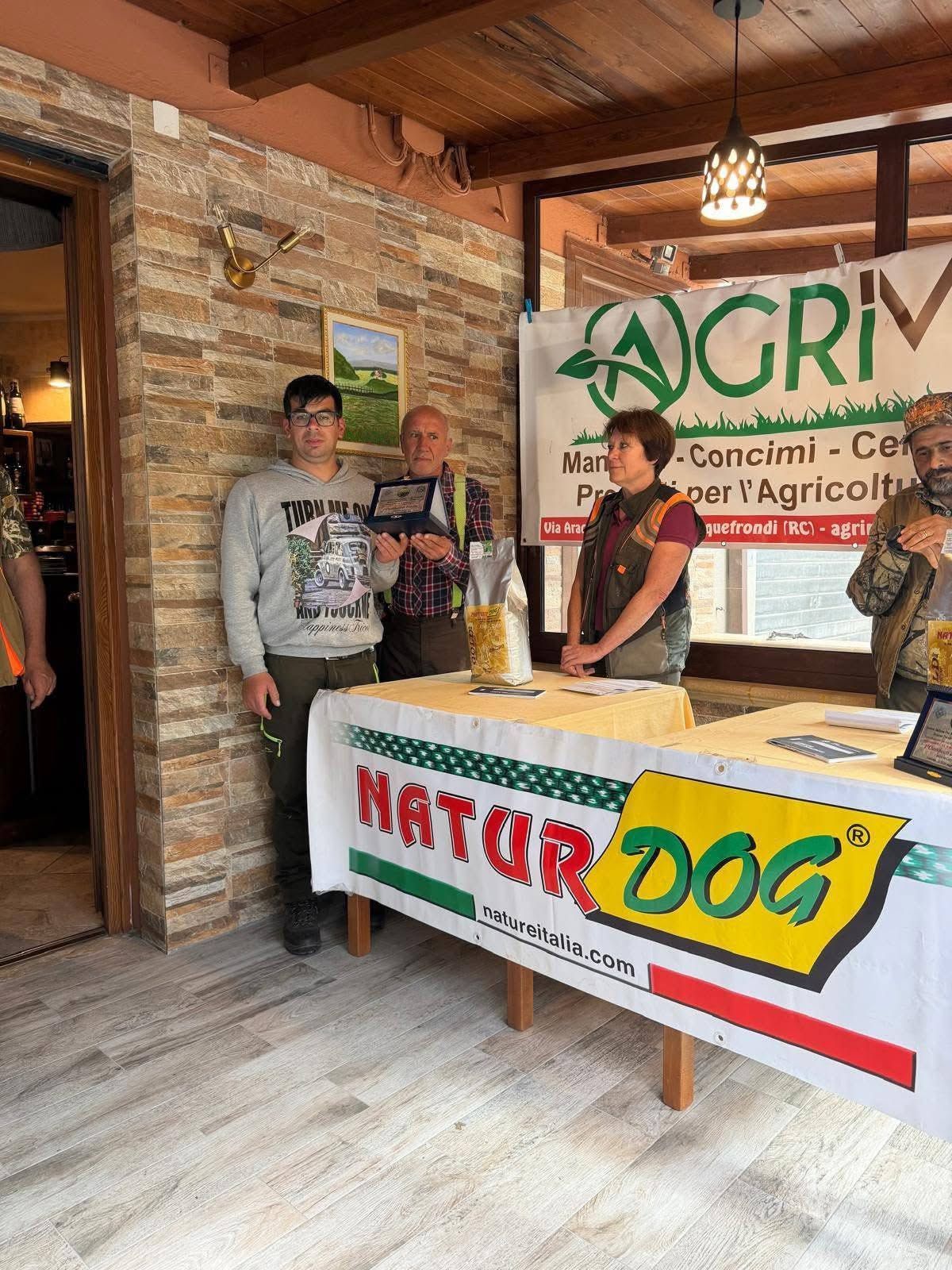 Tre persone dietro un banchetto promozionale di NaturDog in un rustico mercato coperto.