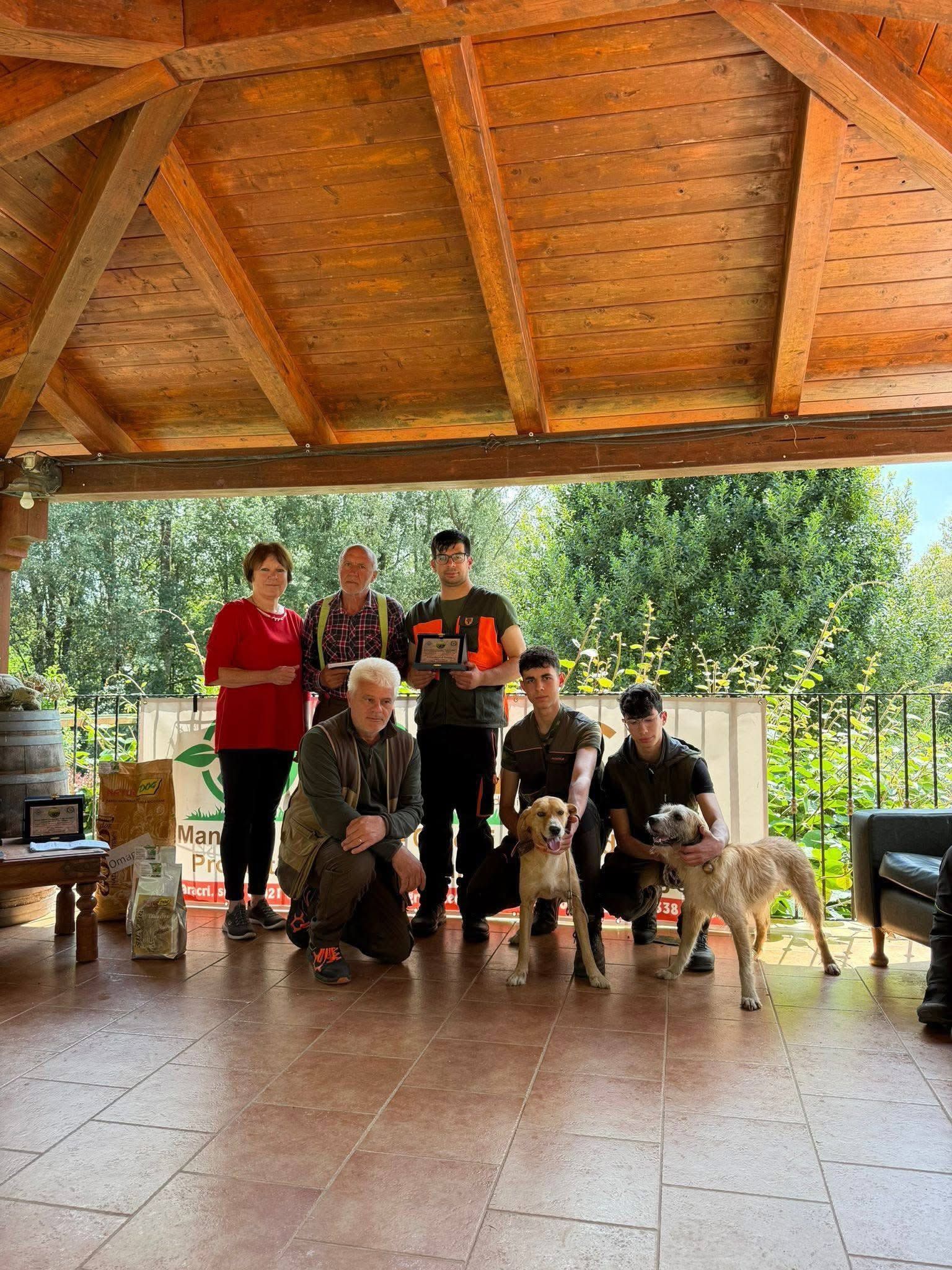 Gruppo di persone in posa con due cani su un patio coperto, con del verde sullo sfondo.