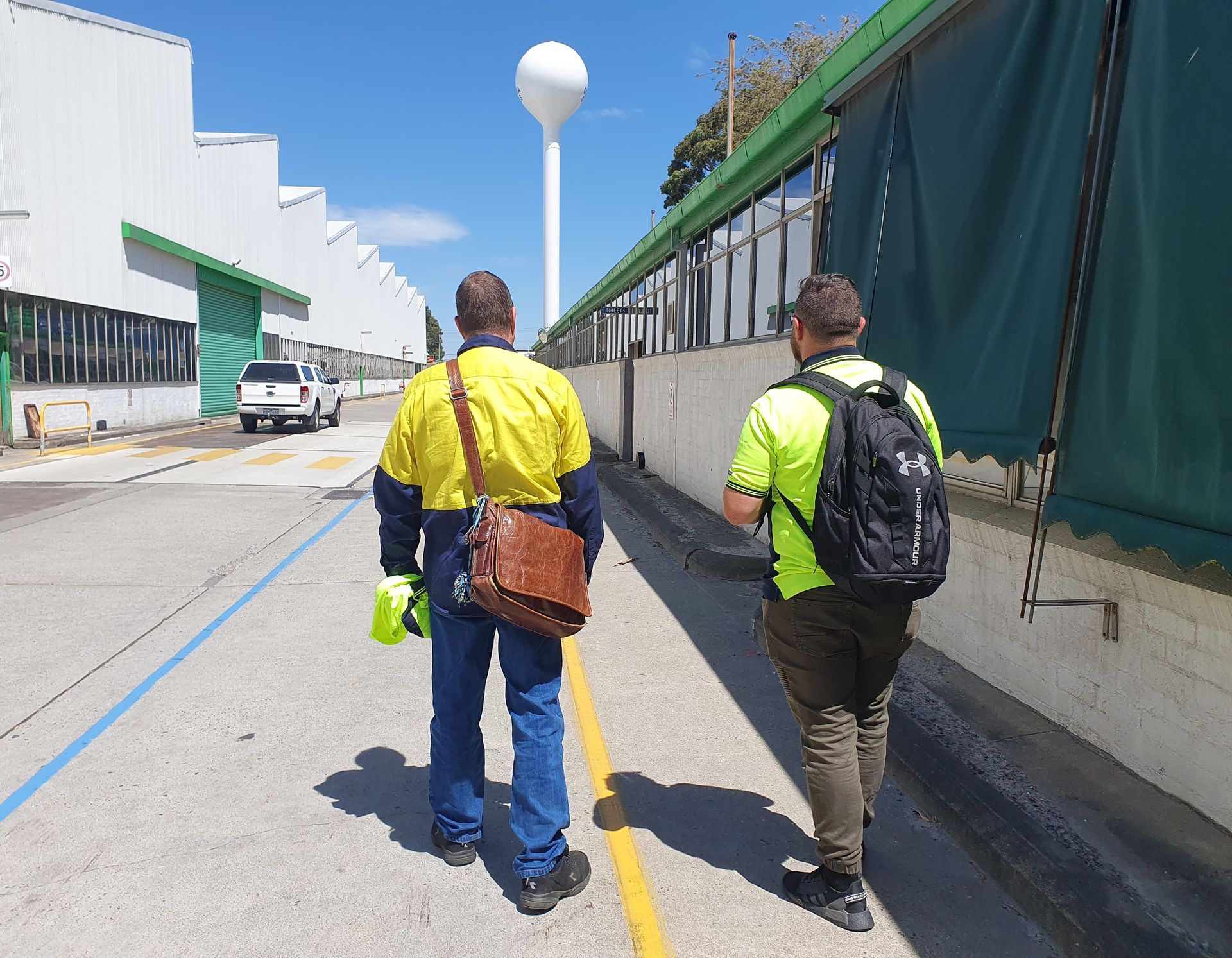 Two people walking along a paved path beside a building and green fence on a sunny day.
