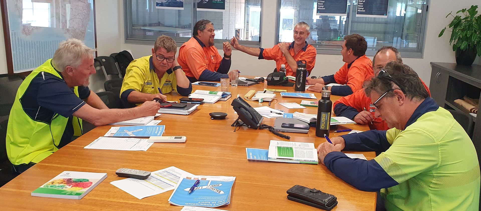 Workers in orange and yellow vests seated around a table reviewing papers in a meeting room