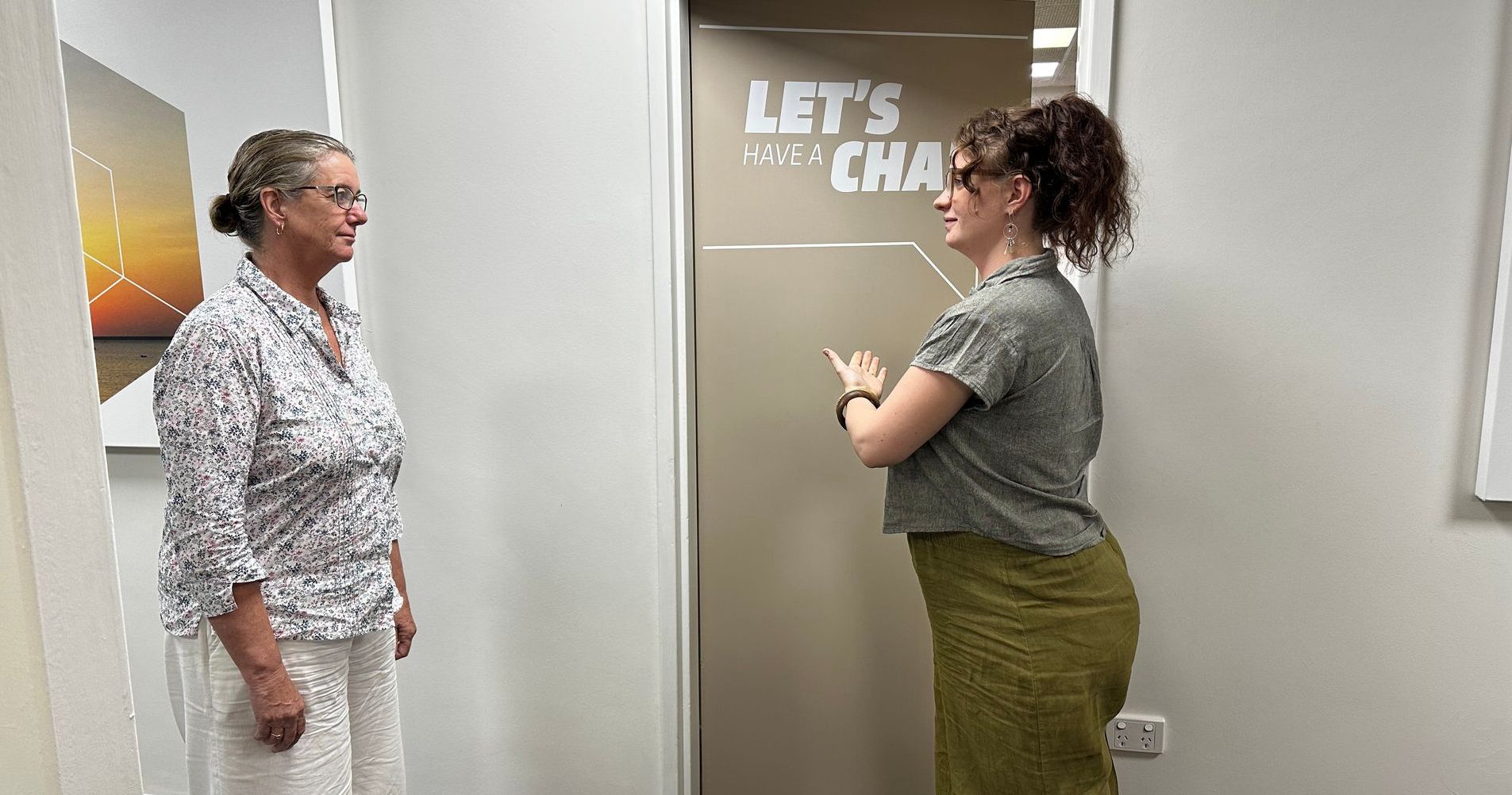 Two women talking in a hallway beside a sign that says “LET’S CHAT.”