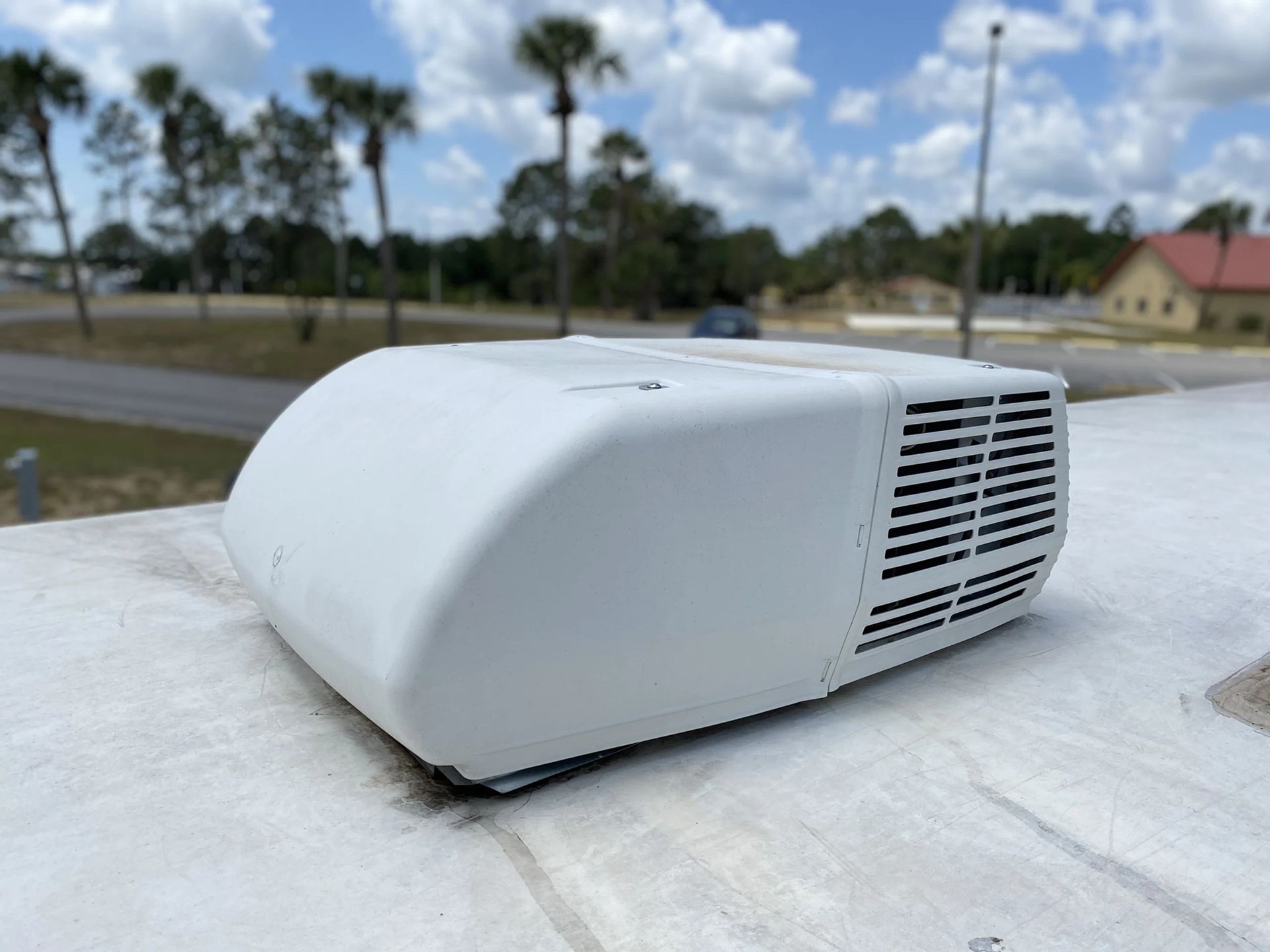 White RV air conditioning unit on a rooftop, with black vents. Background features trees and blue sky.