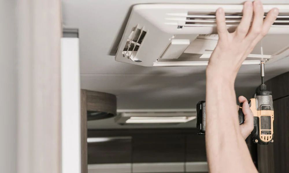 A person is using a power drill to install or repair an RV air conditioner. Interior setting with neutral tones.