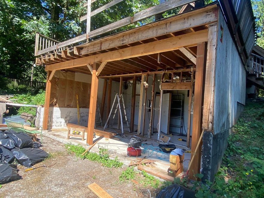 Partially built wooden house with exposed framing, ladder, and construction materials under a deck outdoors