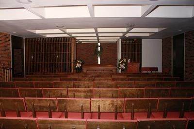 Rows of wooden pews face a brick altar with a central wooden cross in a dimly lit chapel with a coffered ceiling.