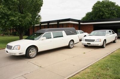 A white hearse and a white stretch limousine parked in a row on a driveway in front of a brick building.