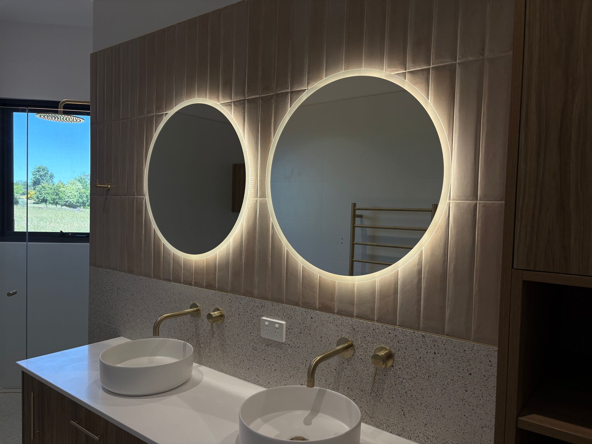 Bathroom with two round, illuminated mirrors above sinks with gold fixtures. Light wood paneling— SJD Electrical Contracting in Orange, NSW