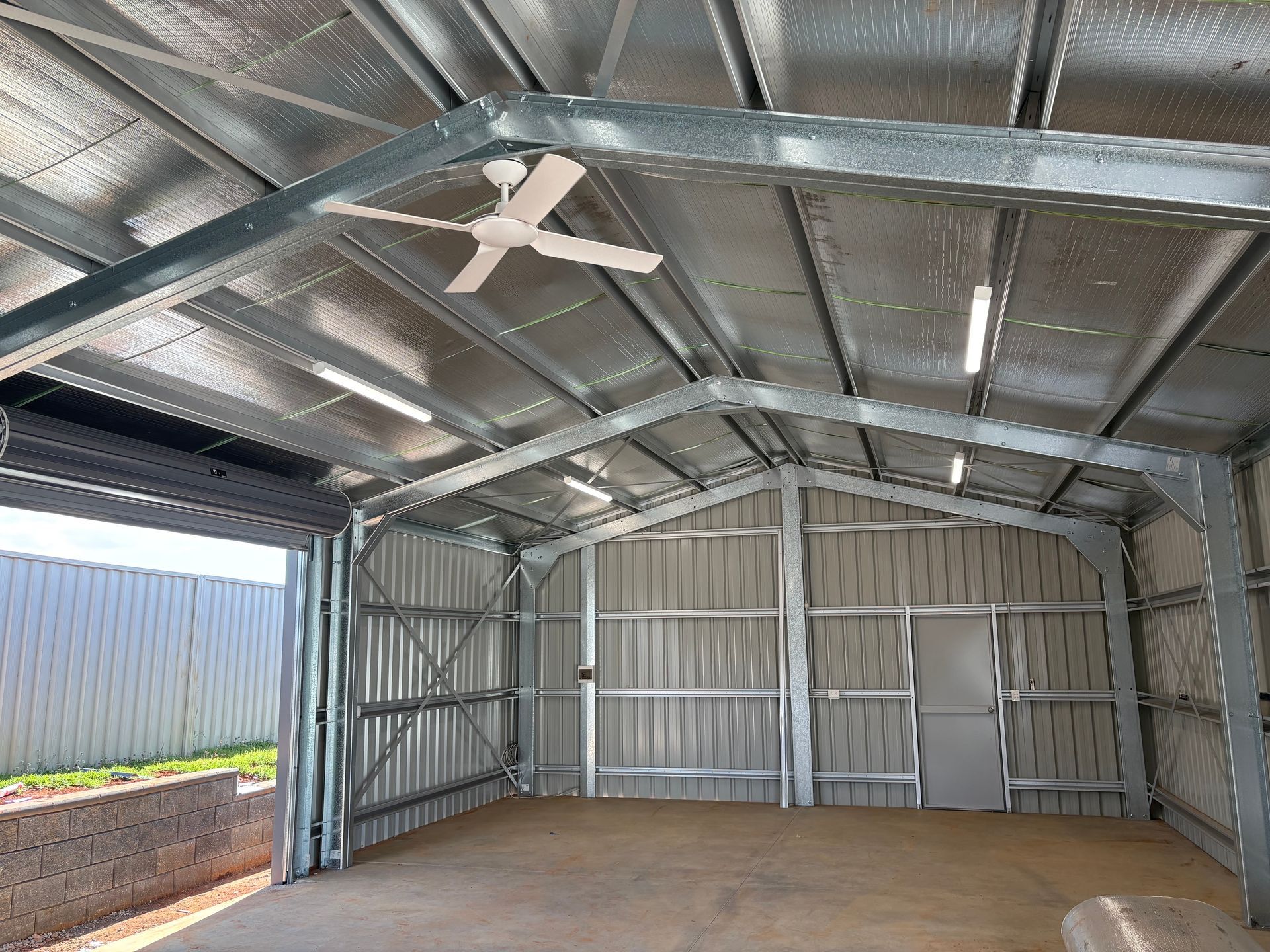Interior of a metal shed with a ceiling fan and lights. Grey walls and floor, empty space.