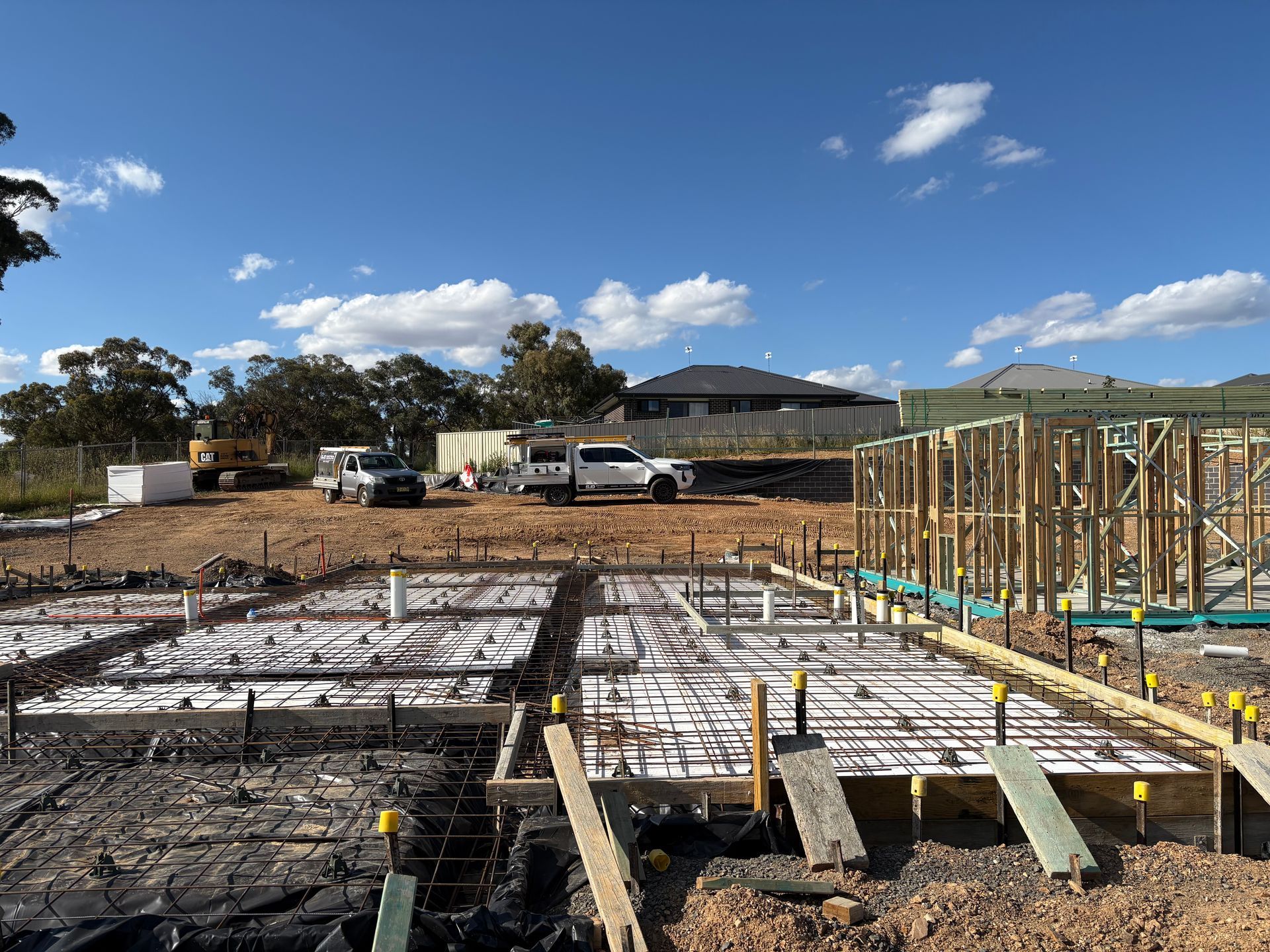 Construction site with a partially built house frame, foundation, and work trucks under a blue sky.