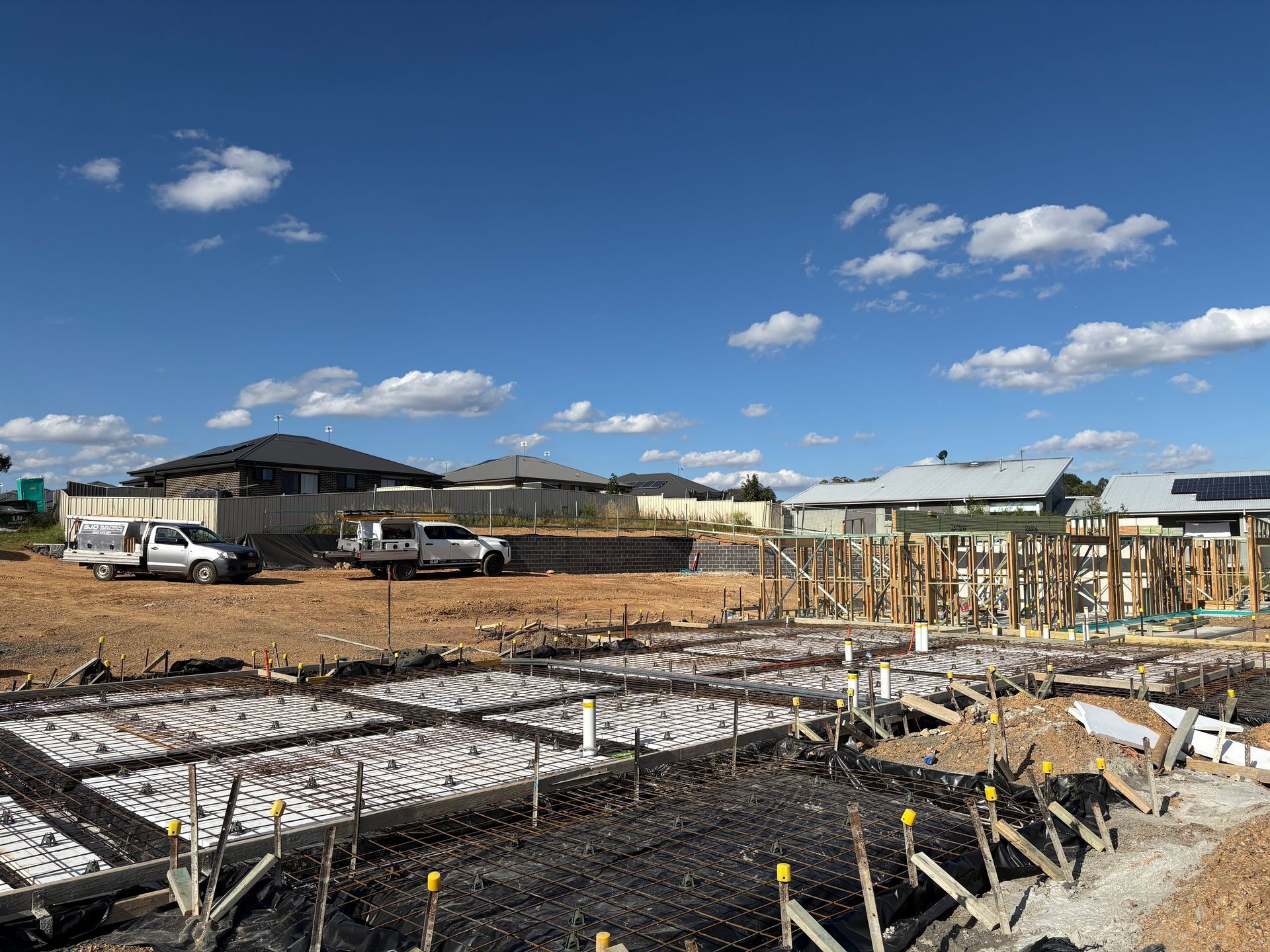Construction site with foundation work. Trucks on dirt, houses in background under blue sky.