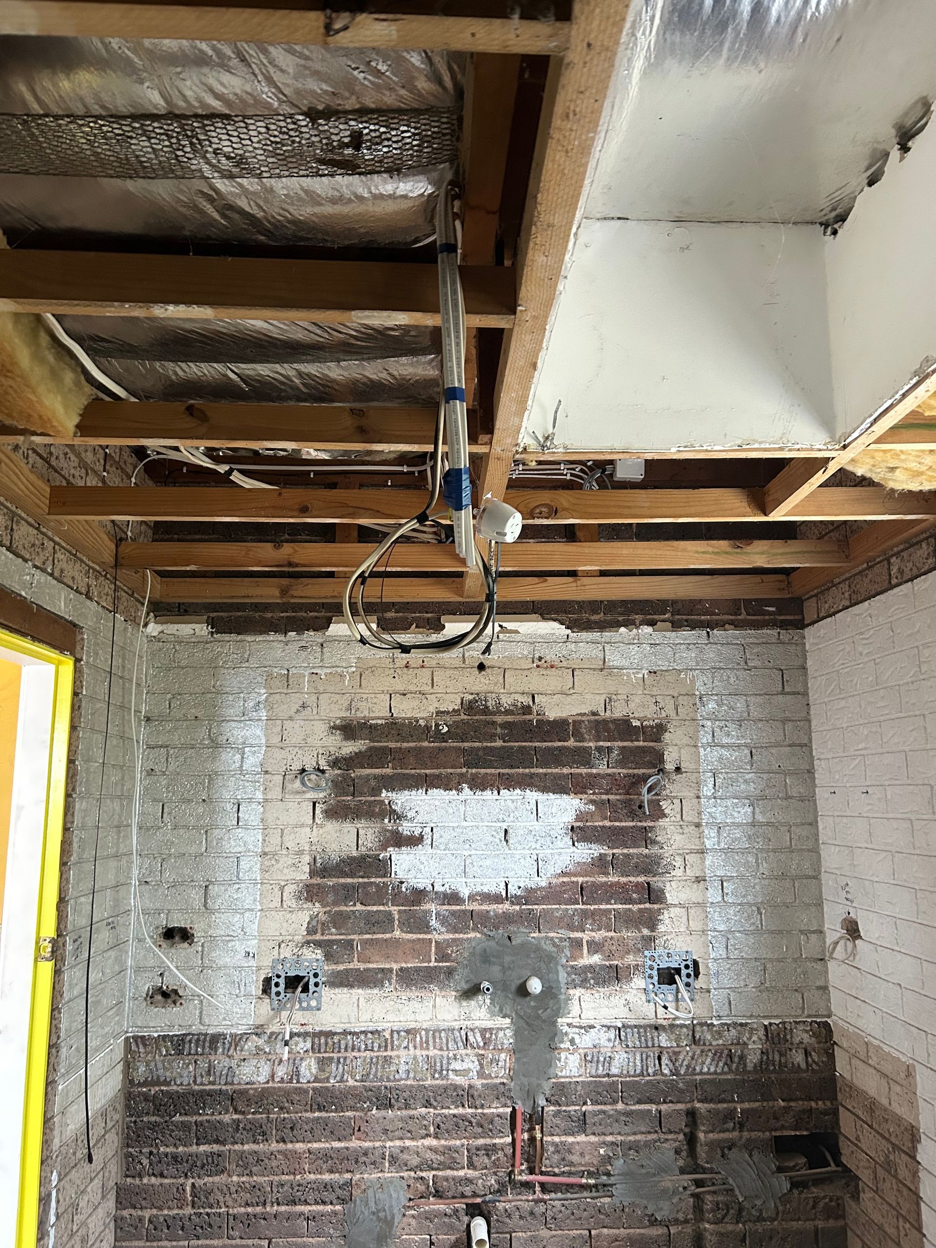 Kitchen under construction, exposed brick wall and wooden ceiling beams, pipes and wires visible.