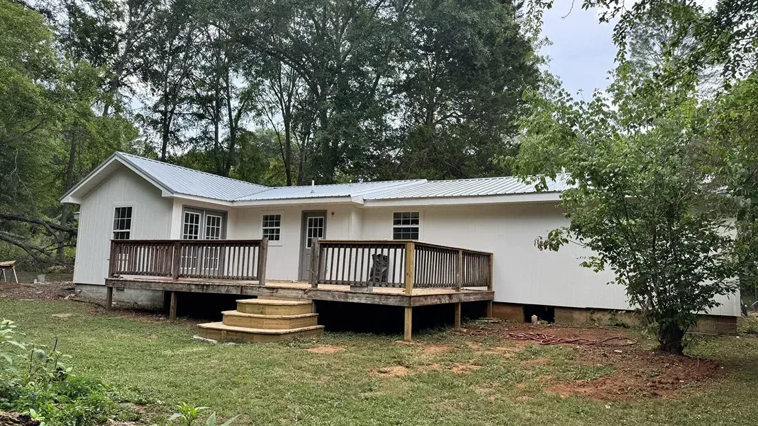 White house with wraparound porch and steps, surrounded by trees and dirt yard