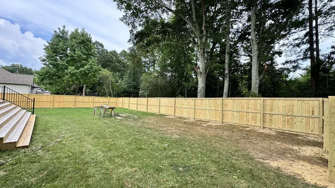 Backyard with a wooden fence, green lawn, and tall trees under a cloudy sky