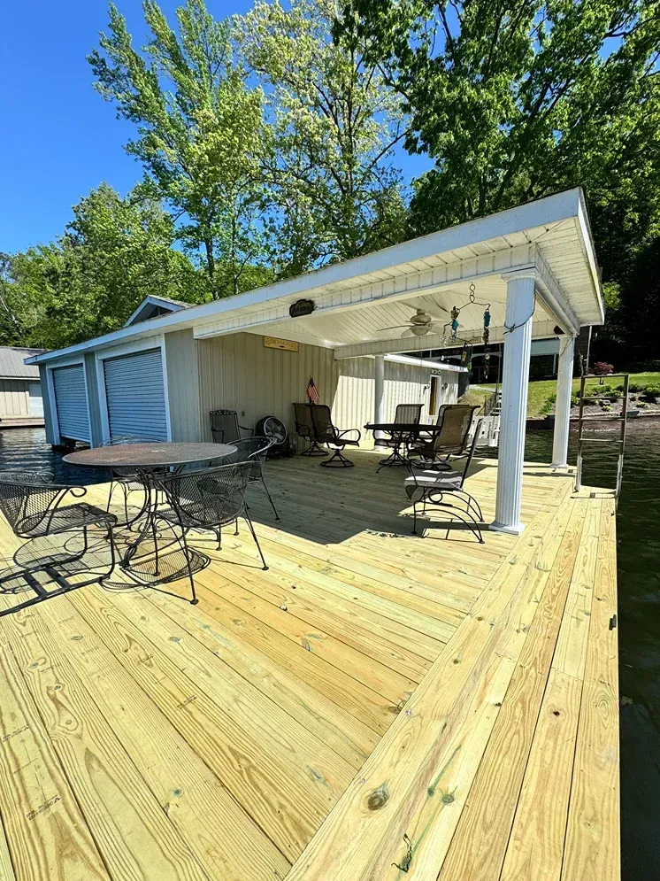 Outdoor wooden deck with patio tables and chairs under a white roof beside a house, trees and blue sky behind