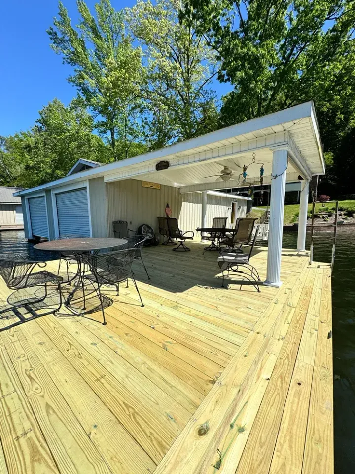 Outdoor wooden deck with patio tables and chairs under a white roof beside a house, trees and blue sky behind