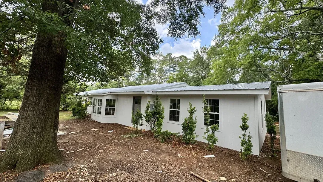 White single-story house with dark windows, surrounded by trees and leaf-covered ground.