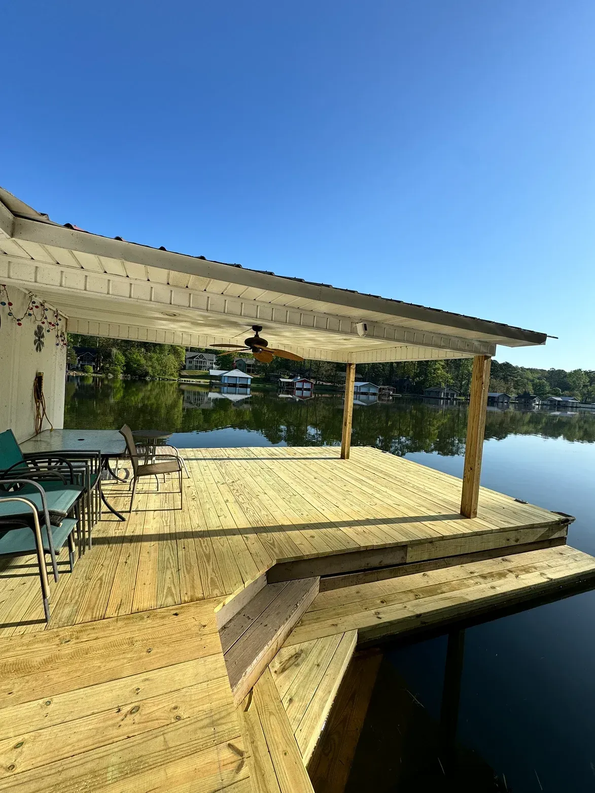 Wooden lakeside deck with covered seating and chairs under a bright blue sky, overlooking calm water
